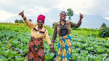 Two women hold hands and lift their free hands into the air with thumb's up. They are standing in front of a green field of vegetables with a mountian in the far distance.  