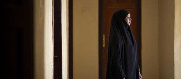 Photograph of a woman in a black abaya standing in a doorway along a sunlit corridor.