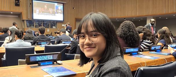 Young woman seated in an auditorium-style conference room with attendees seated at wooden desks, presentation on screen.