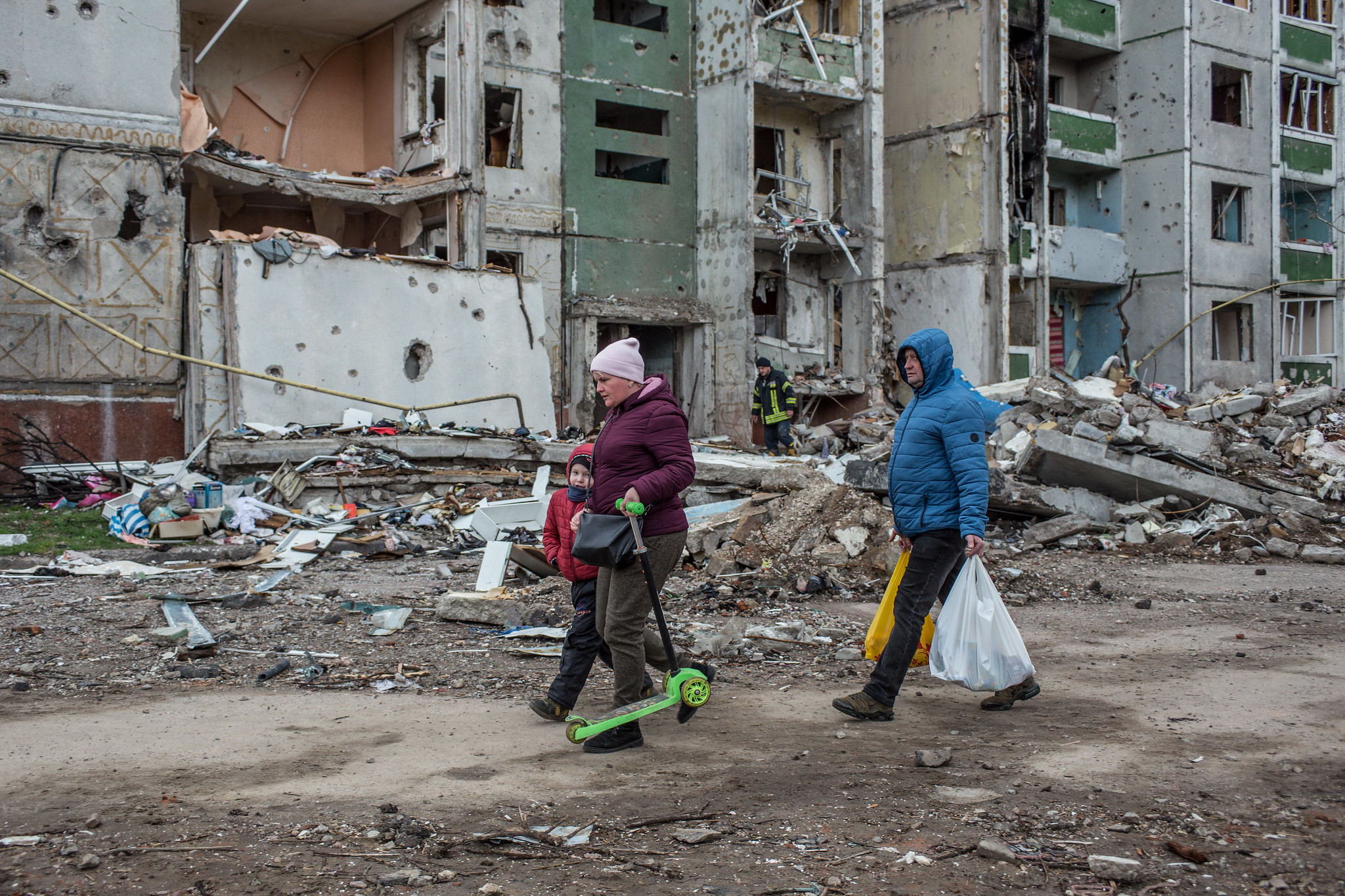 A family walks down a street with damaged buildings in the background.