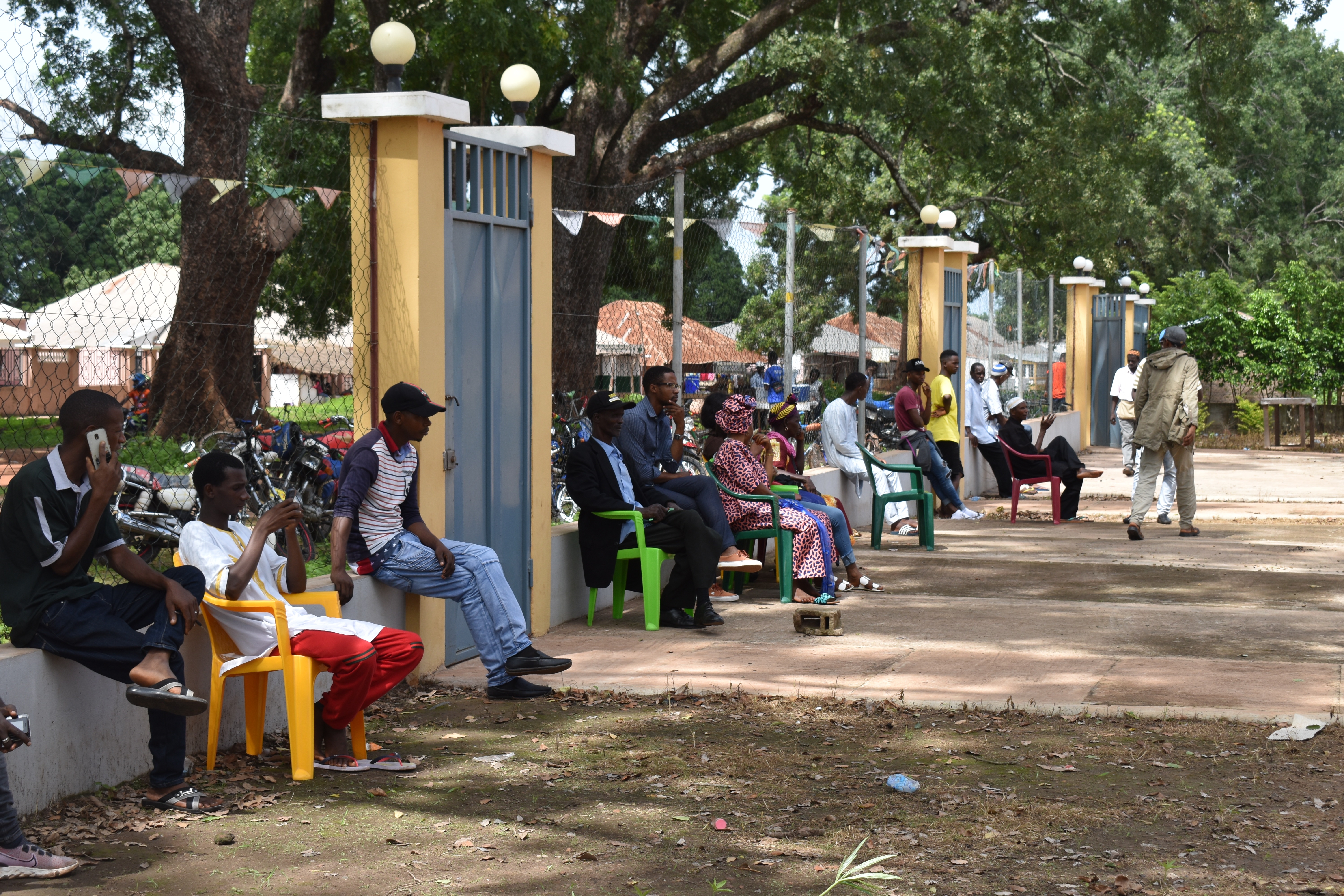 Pessoas na fila trazem suas próprias cadeiras plásticas ou fazem uso do muro da propriedade por falta de assentos na Casa de Justiça, em Gabu