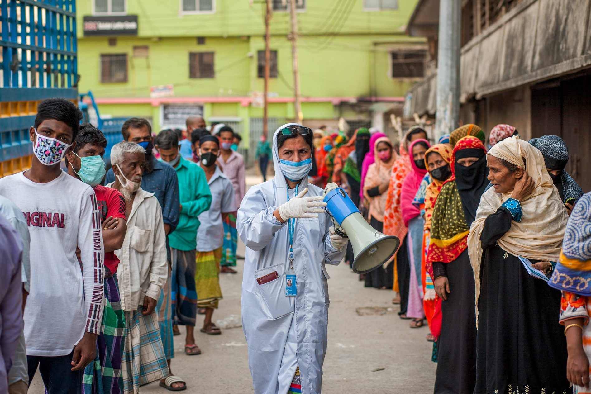 Photo of a woman wearing COVID-19 protective clothing, holding a loudspeaker walking along queuing people in Bangladesh.