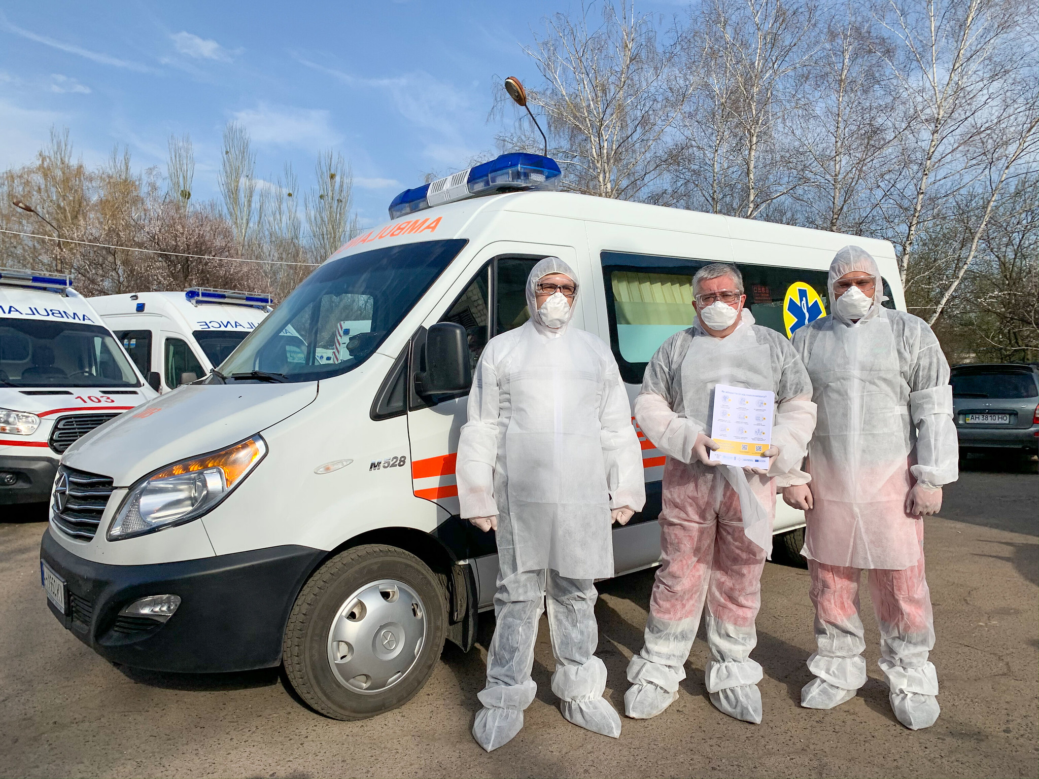 Medical workers in protective equipment stand in front of an ambulance.