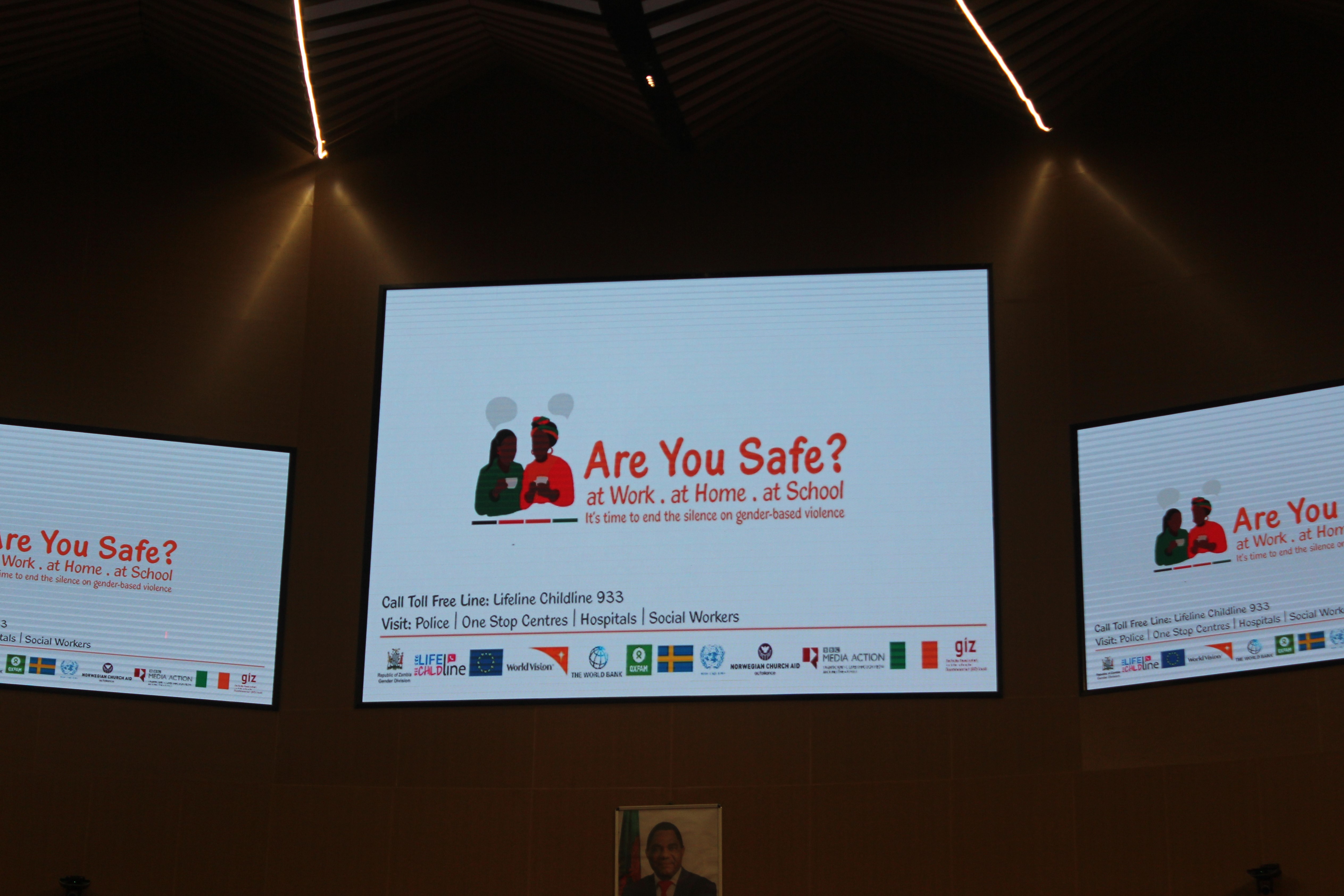 A screen at the TV launch of 16 Days of Activism Campaign in Zambia. The text reads: "Are You Safe? at Work. at Home. at School. It's time to end the silence on gender-based violence" This is part of the local theme for 2022