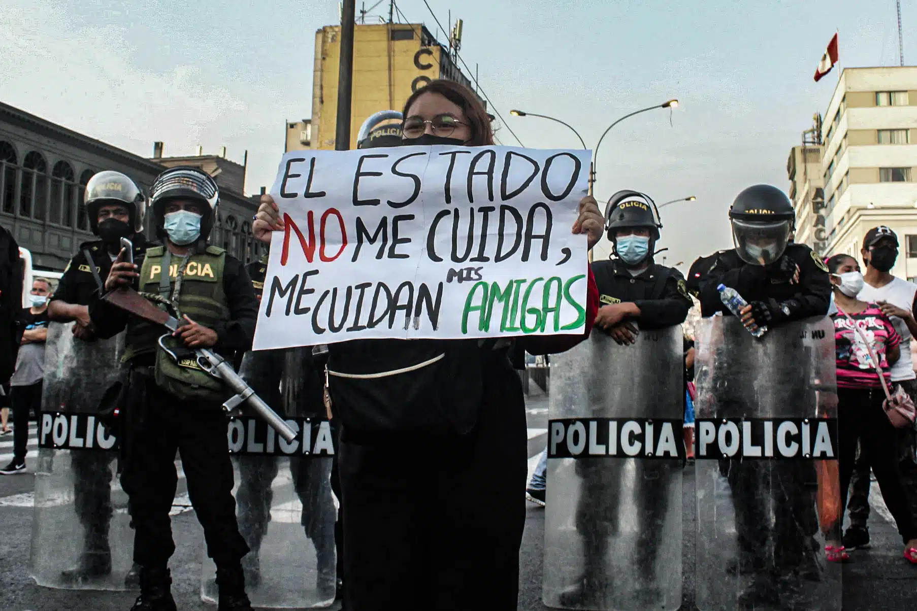 Foto de una manifestación. Una mujer lleva un cartel que dice "El Estado no me cuida, me cuidan mis amigas". Atrás de ella hay policías con cascos y escudos.