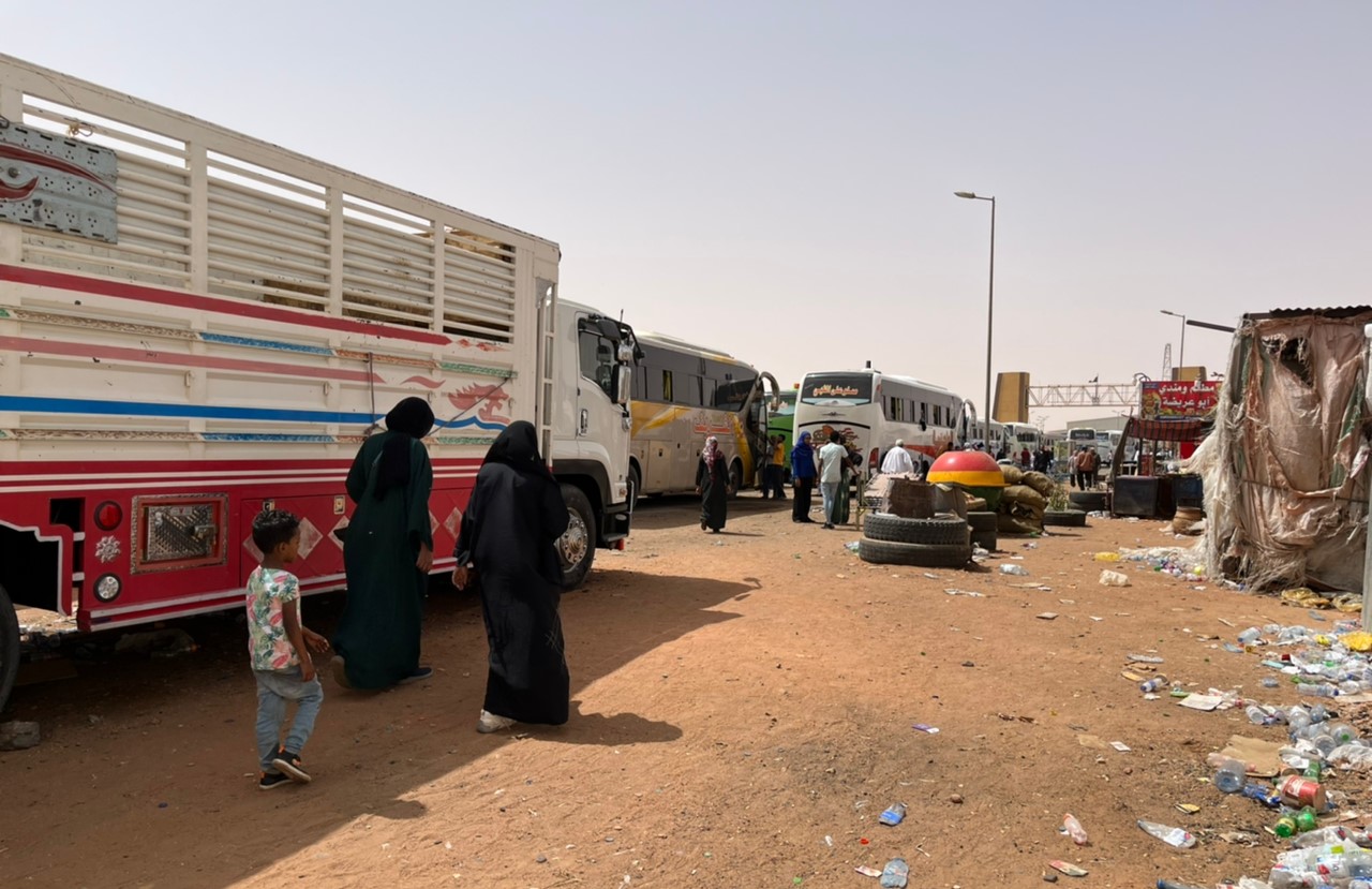 Two women and child walking next to bus and truck in line