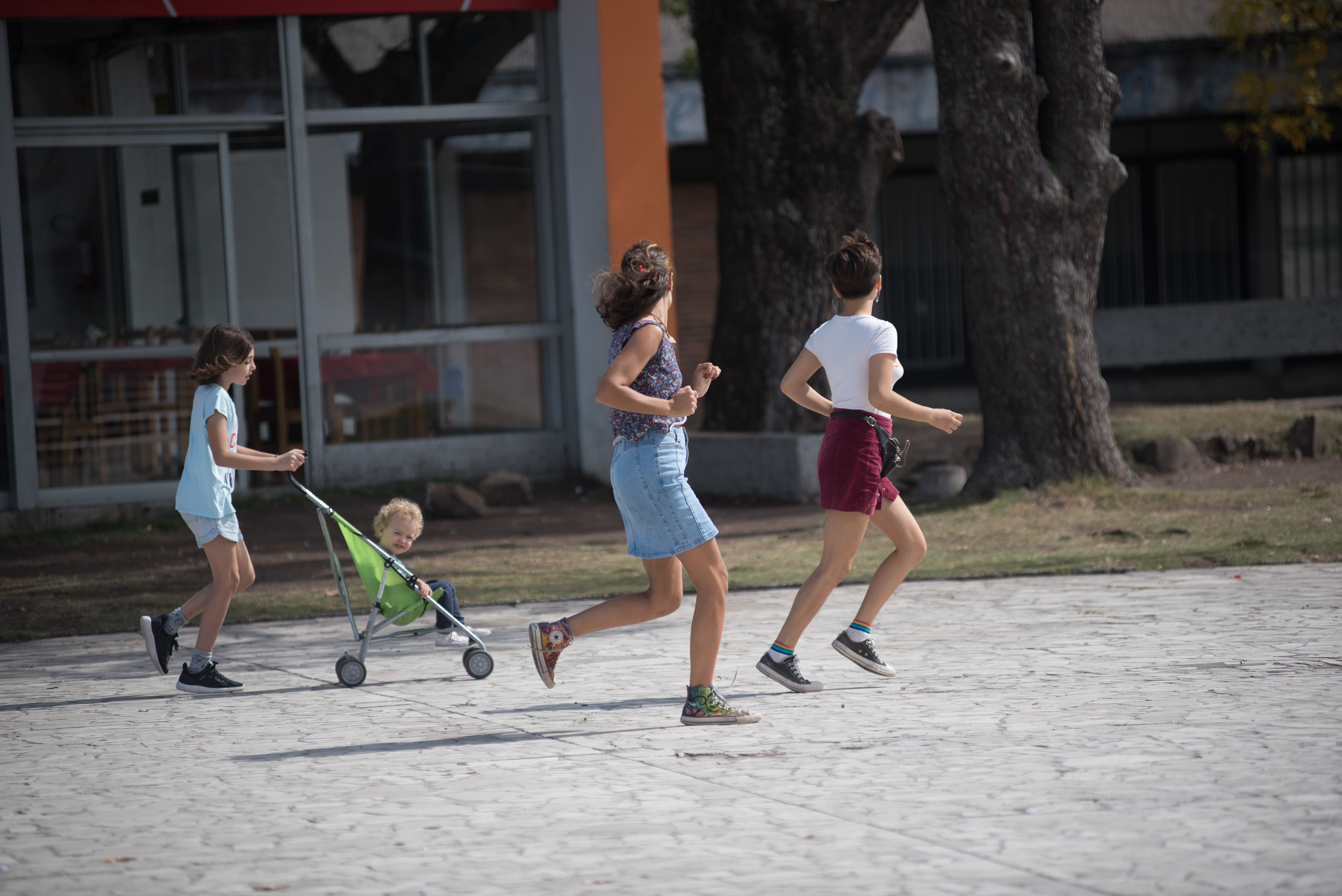 Mujer y dos niñas corren y juegan en plaza de Montevideo, una de ellas traslada a un bebé en un coche.