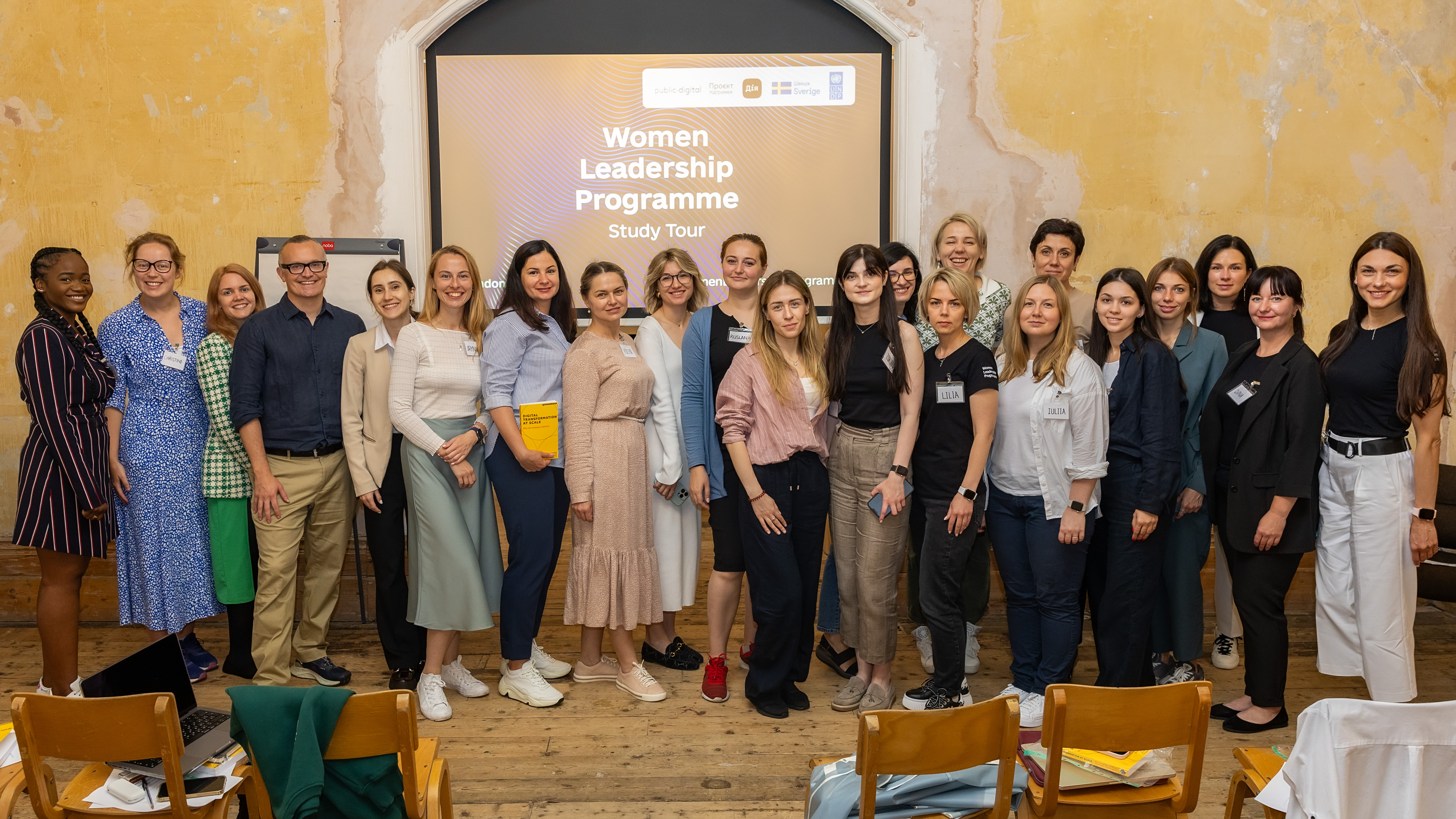 A group of people (mostly women but also a few men) are standing in front of a big screen with the inscription "Women Leadership Programme"