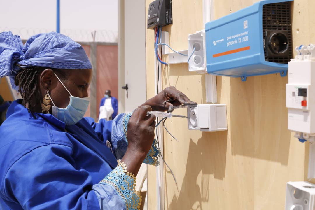 Una mujer vestida de azul y una mascarilla, reparando un dispositivo eléctrico de pared
