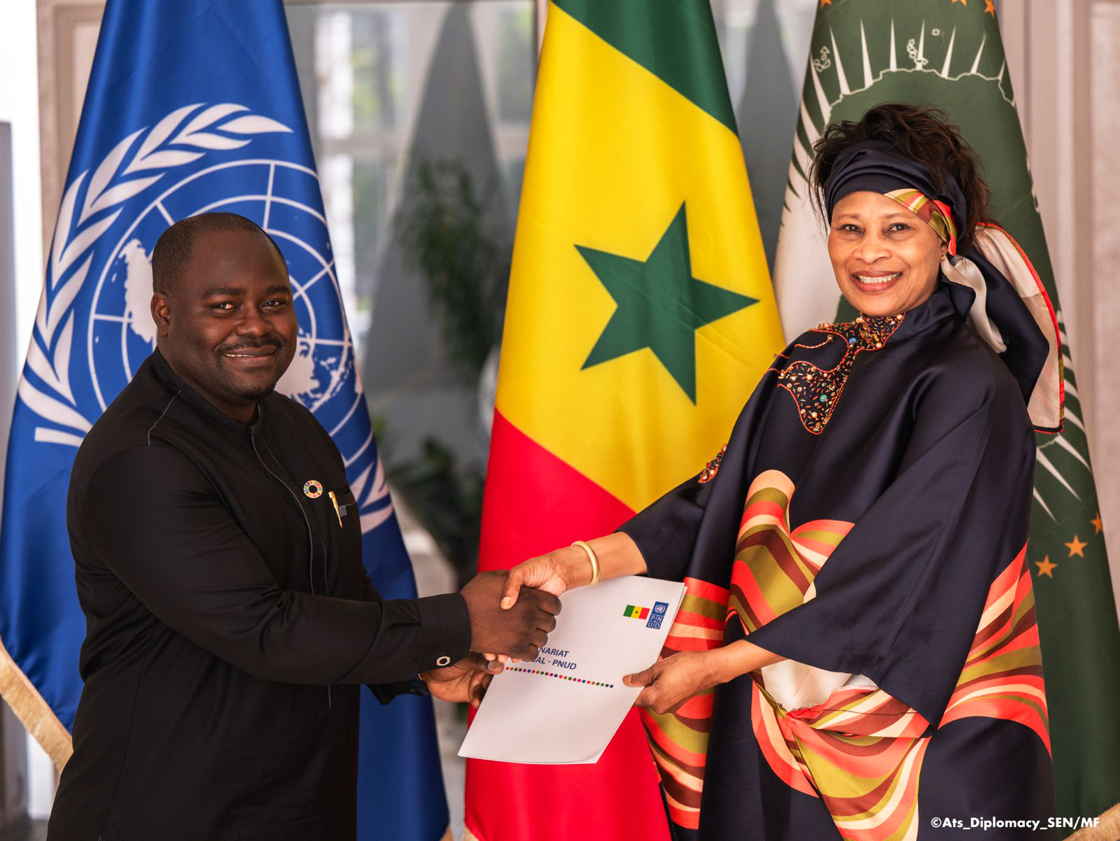 un homme et une femme vêtus de noir, l'homme donne un porte document à la femme, serrière eux les trois drapeaux des Nations Unies, du Sénégal et de l'Union africaineau du 