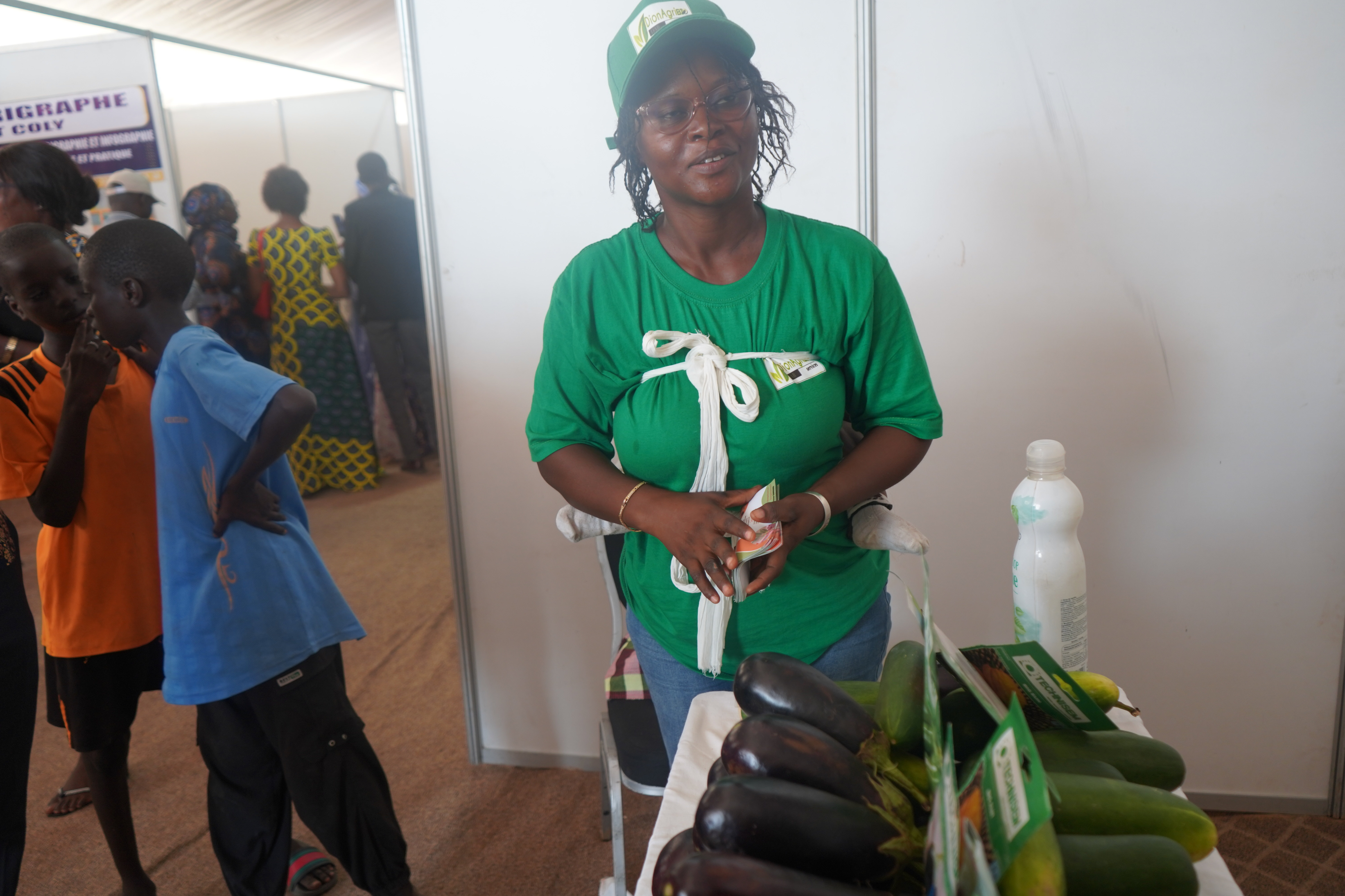 jeune femme en t-shirt vert avec un bébé sur le dos, debout devant des légumes