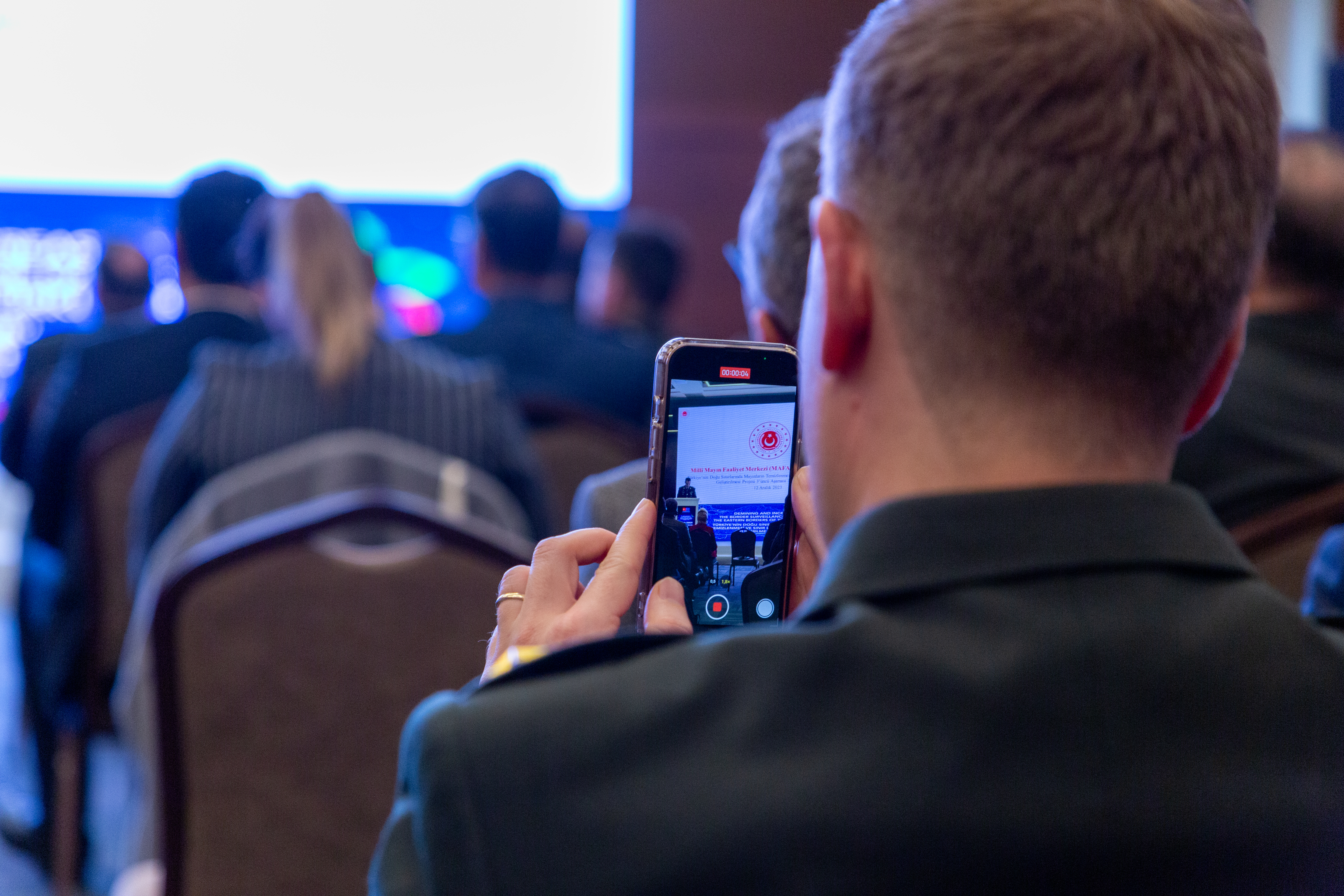 People in a conference room, camera focused on a persons cellphone taking a photo of the stage