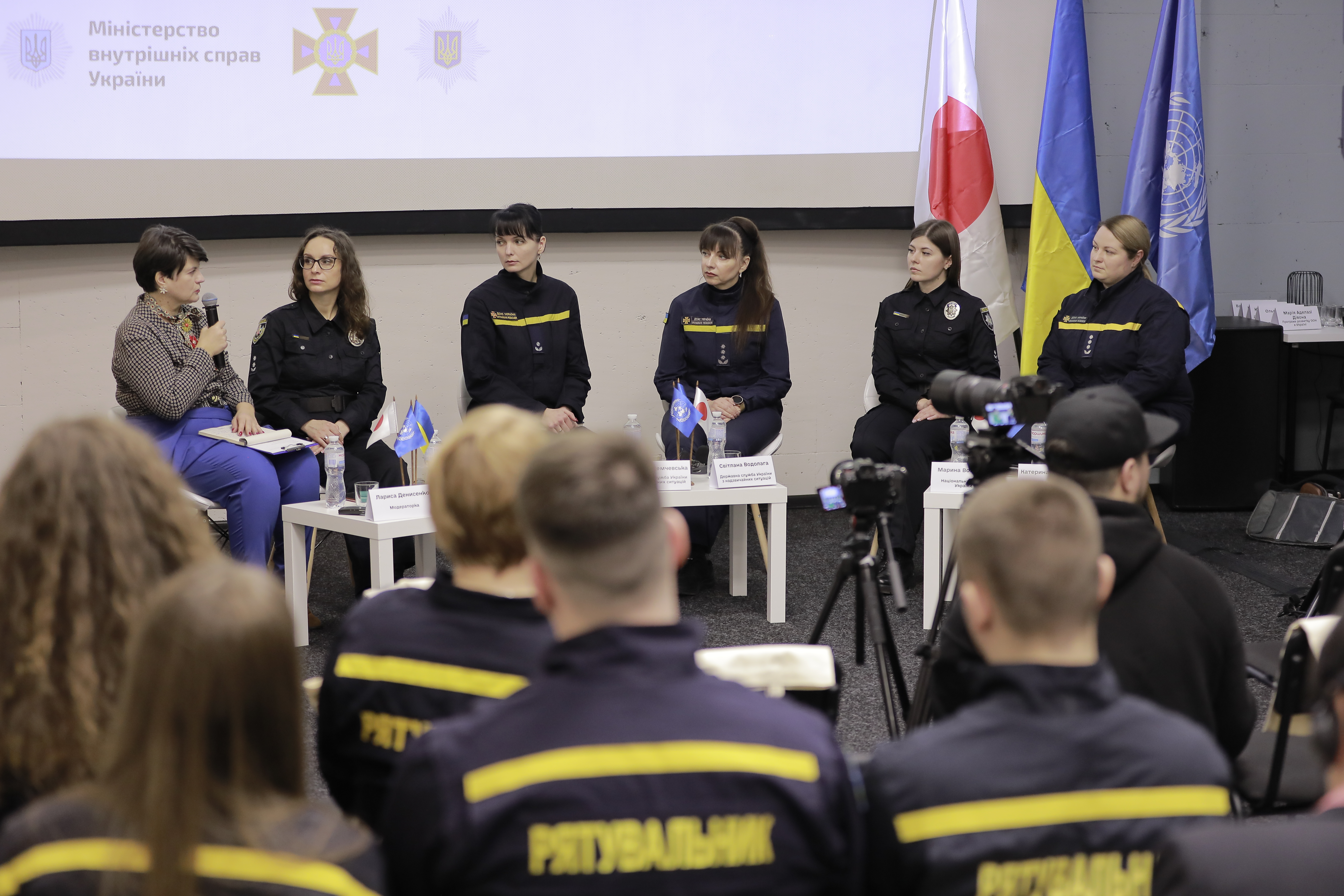 A panel discussion is taking place in a room with a group of women seated in front of an audience. The panelists, some of whom are dressed in black uniforms with yellow trim, are sitting behind a table with name tags and microphones. There are national flags of Ukraine, Japan and the United Nations behind them. In the foreground, the back of the audience is visible, mostly consisting of individuals wearing jackets with "Rescuer" inscription.