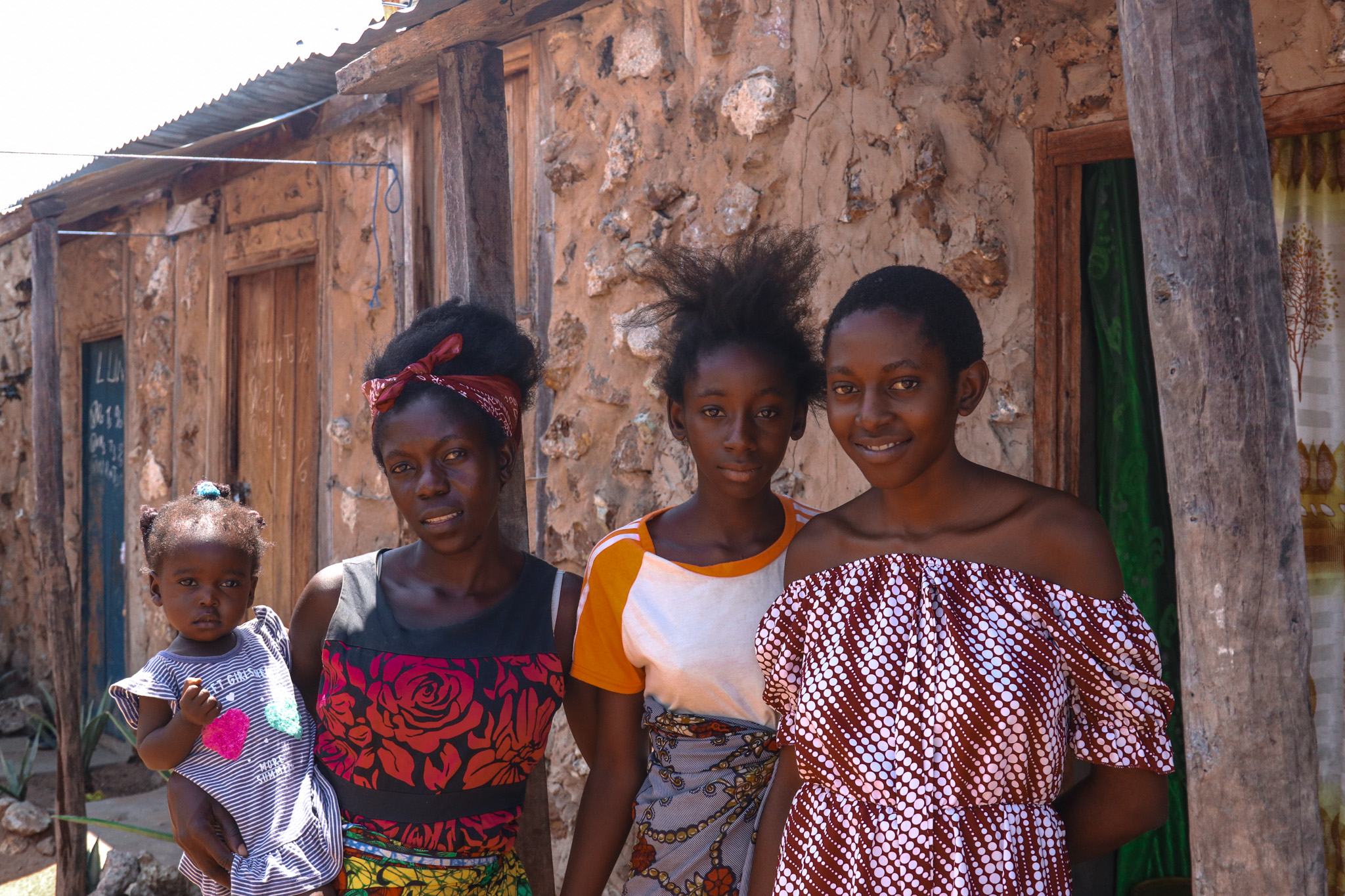 An image of Nalucha holding her daughter, a little girl, in her arms. She flanked by her sisters, in front of her home