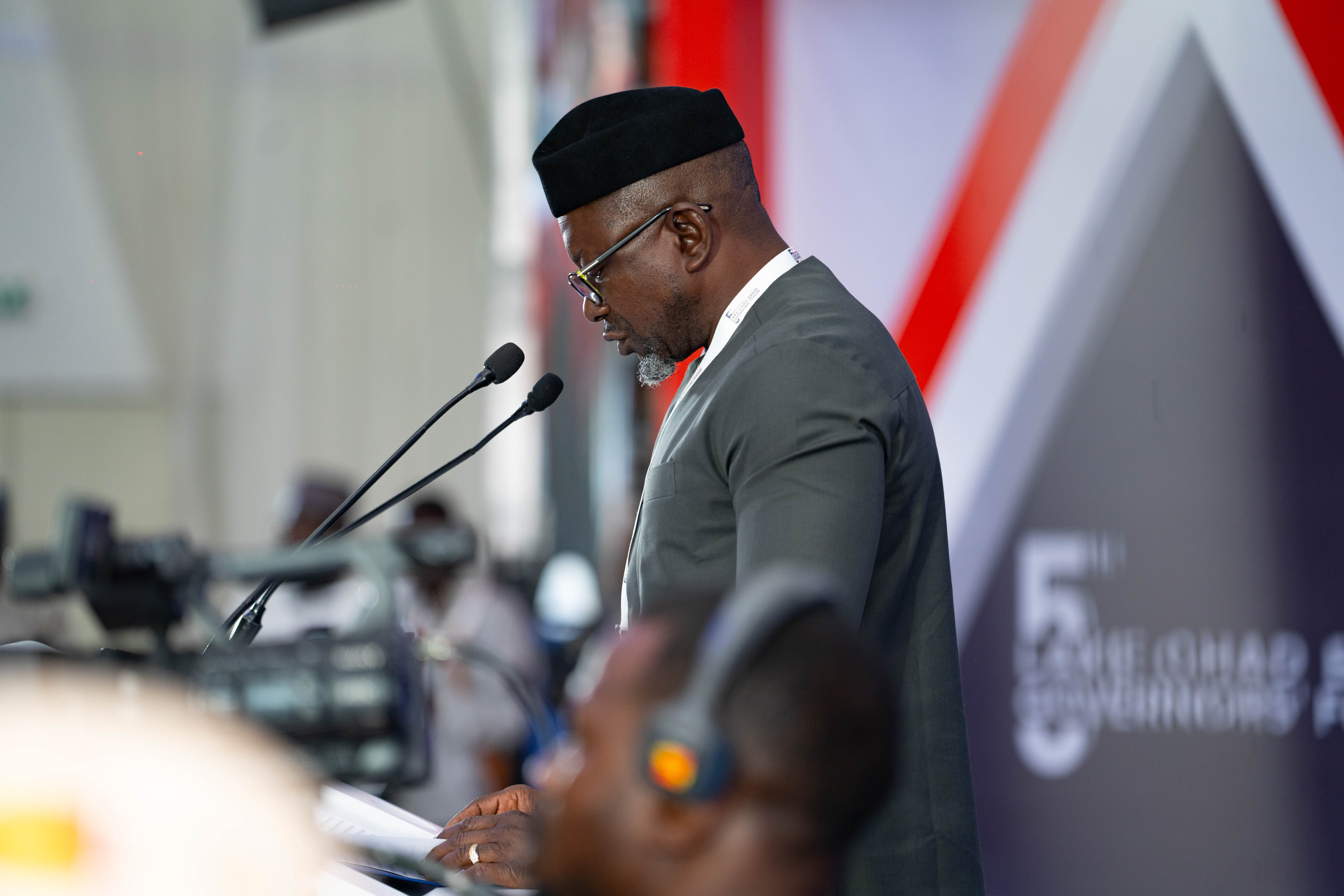 A man in formal attire speaks at a podium, with microphones and an audience in the background.