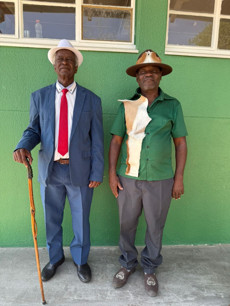 Two men stand side by side against a green wall. One wears a suit and a hat, the other a green shirt with a patterned accent.