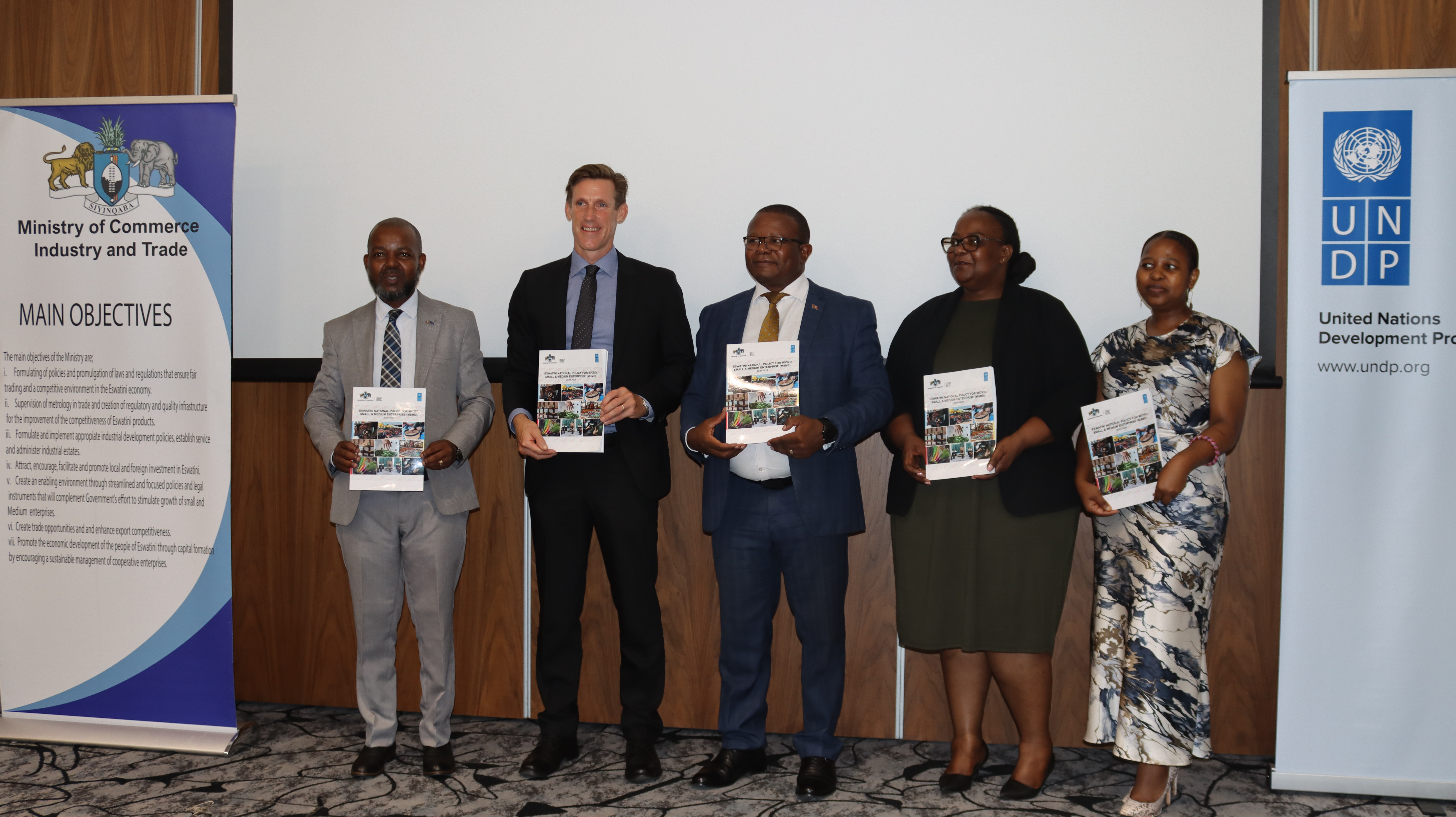 Five people, dressed in formal attire, holding documents, stand together in a conference setting.