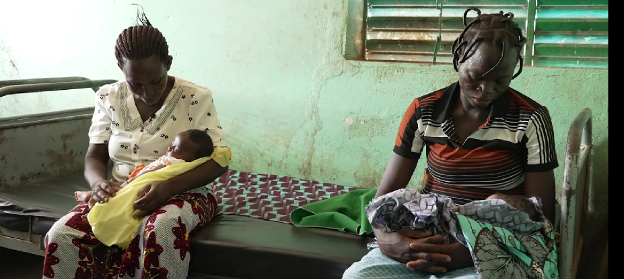 Two women sitting on a bed, each holding a baby in a simple, dimly lit room.