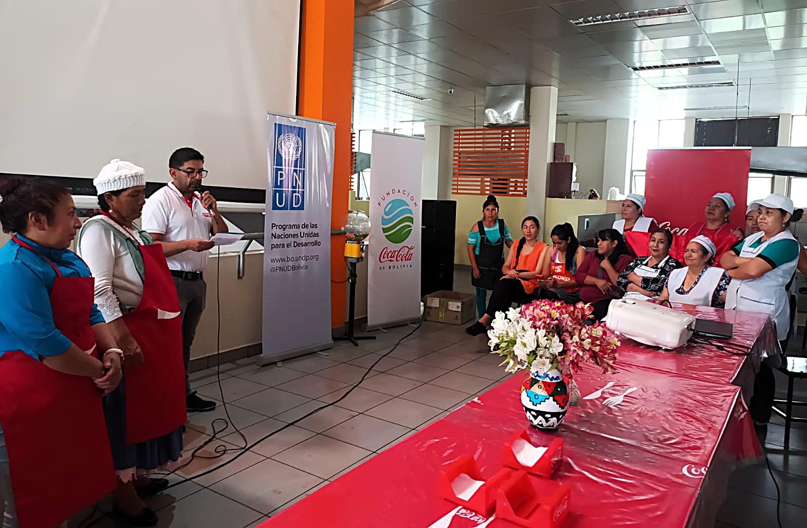 Group of people in an indoor setting, some speaking at a table with flowers and decorations.