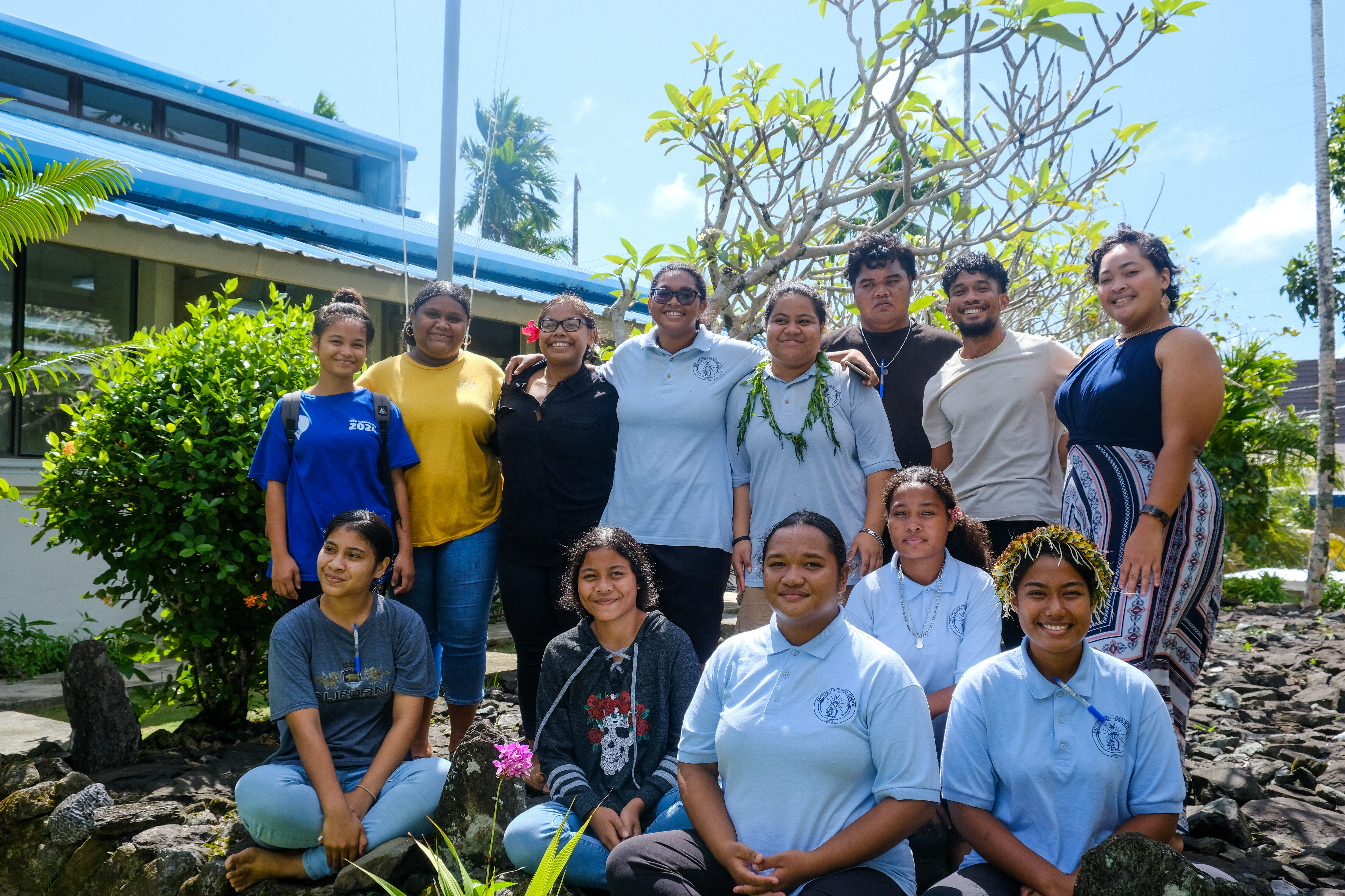 Group of diverse individuals posing outdoors, surrounded by greenery and a sunny sky.