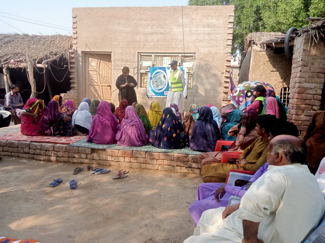 A group of people sitting on the ground, listening to a speaker in a rural setting.