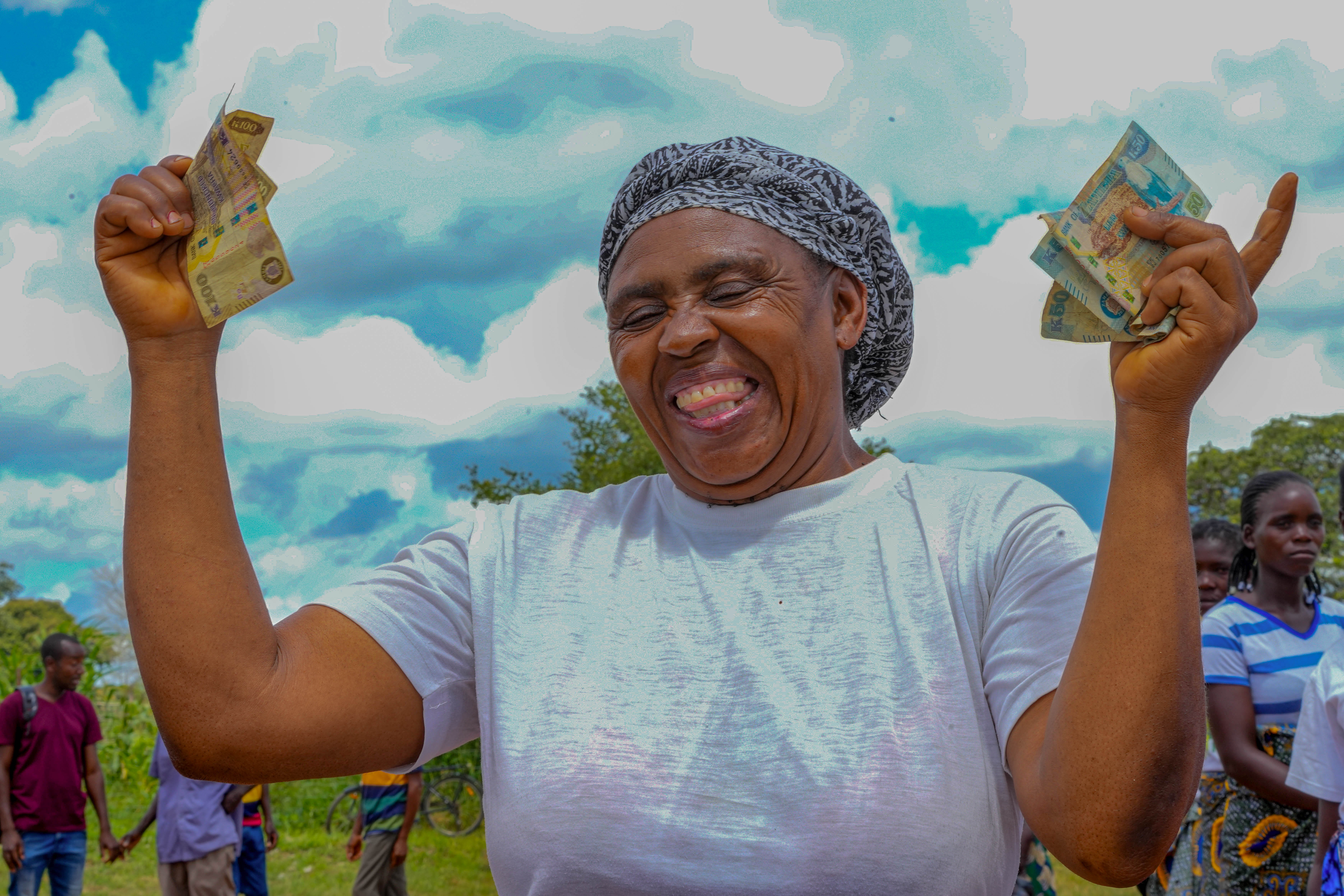 A joyful woman with arms raised, holding cash, smiles against a blue sky with clouds.