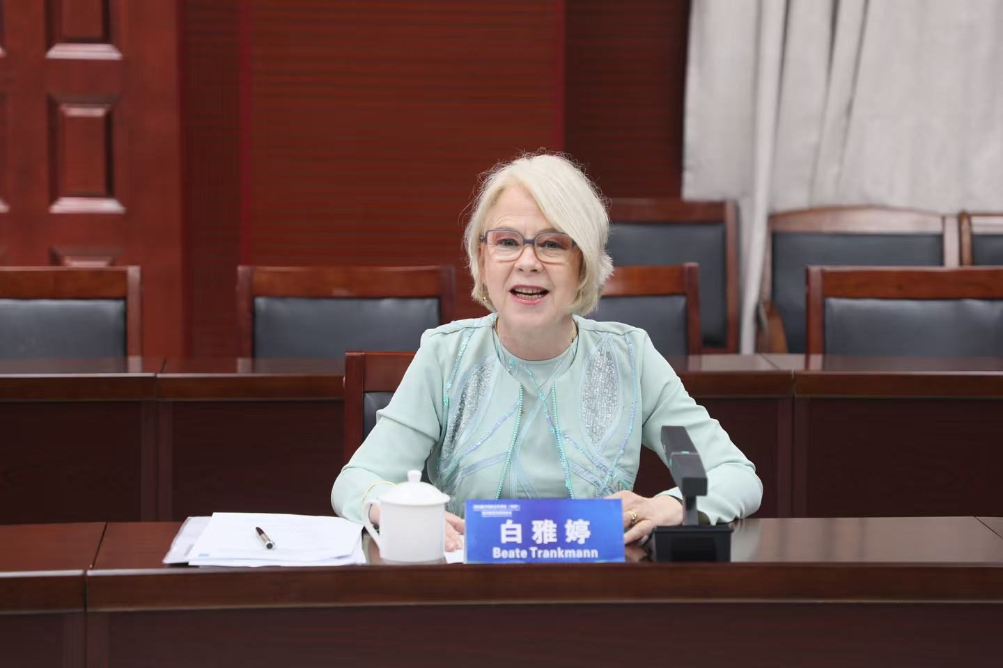 A woman with glasses and white hair smiles while sitting at a conference table.