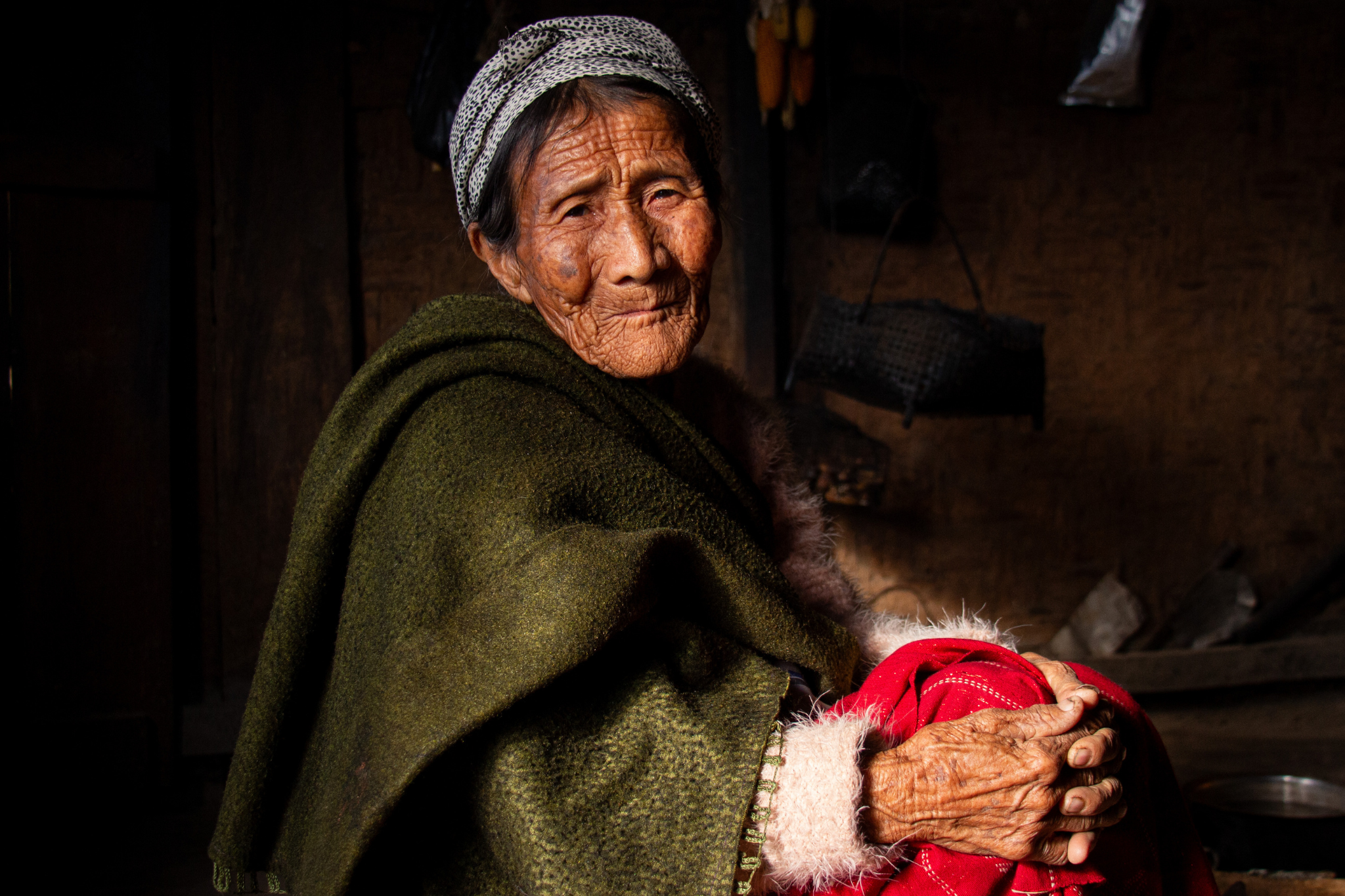 Elderly woman in a green shawl, smiling warmly while holding a red garment in a dimly lit room.