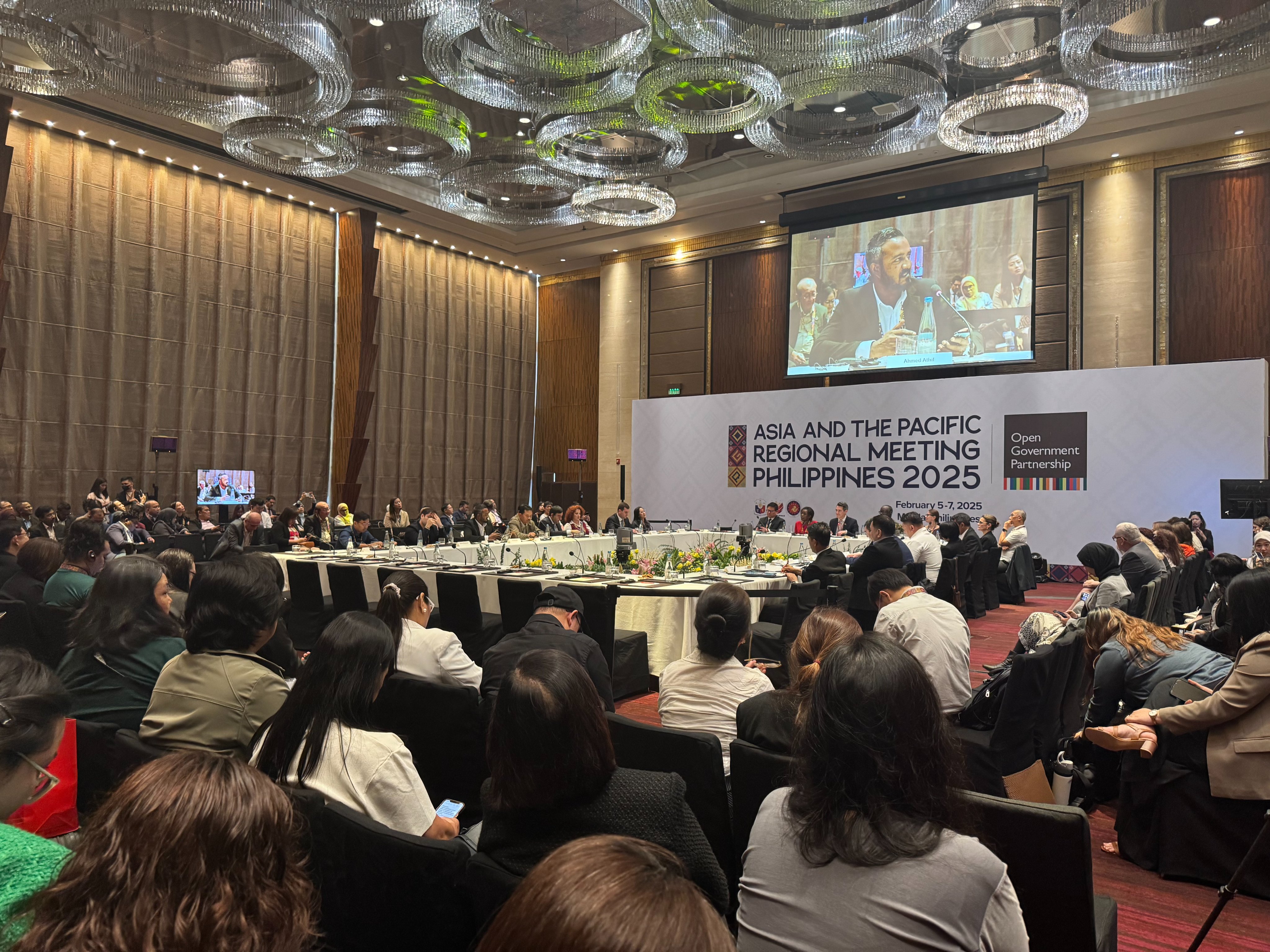 A conference setting with attendees seated at round tables, listening to a speaker on stage.