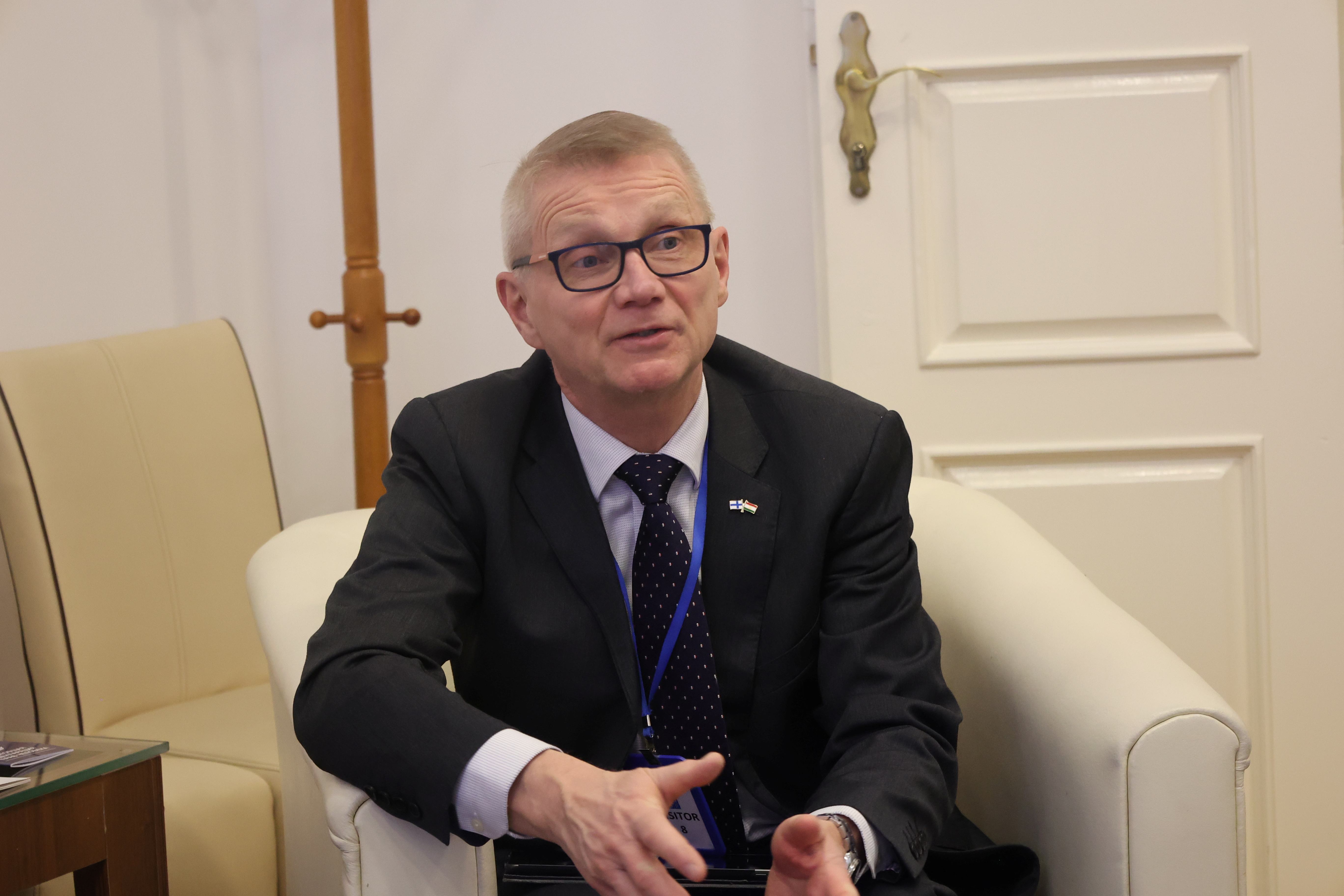 A man in a suit sits on a chair, gesturing while speaking in a formal setting.