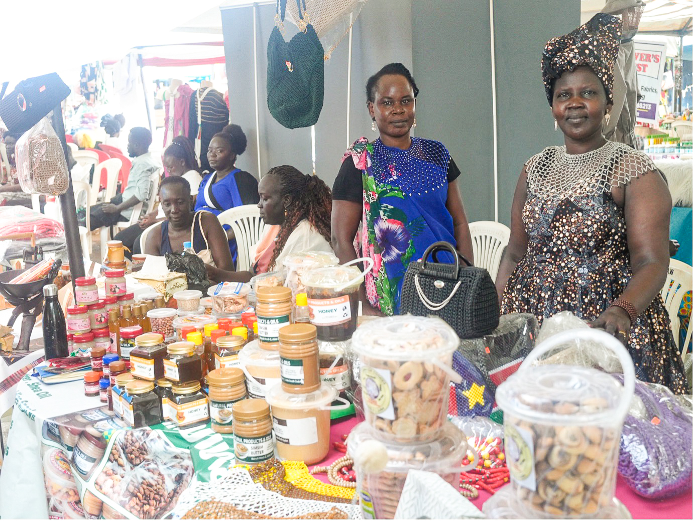 Two women smile behind a vibrant market stall filled with jars and bags of various products.