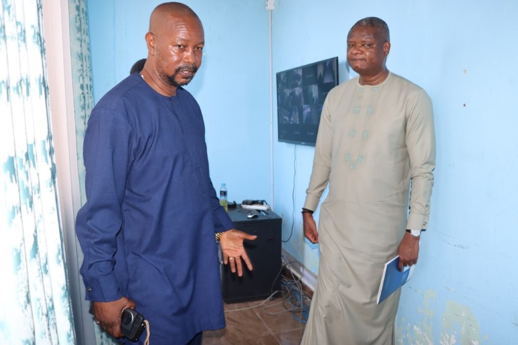 Two men in traditional attire conversing indoors, with a blue wall and a television in the background.