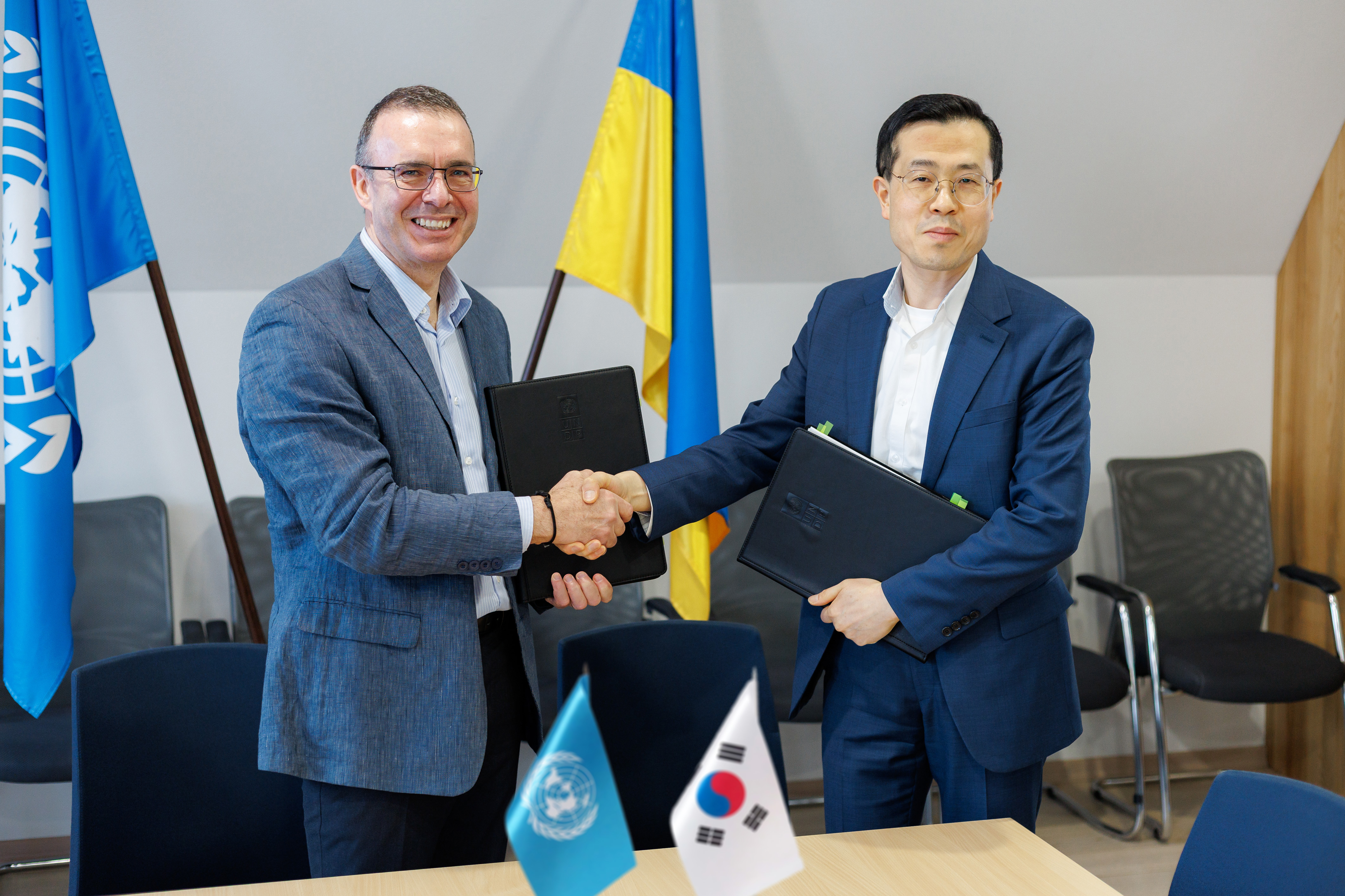 Two men shake hands at a table, with Ukrainian and South Korean flags in the foreground.