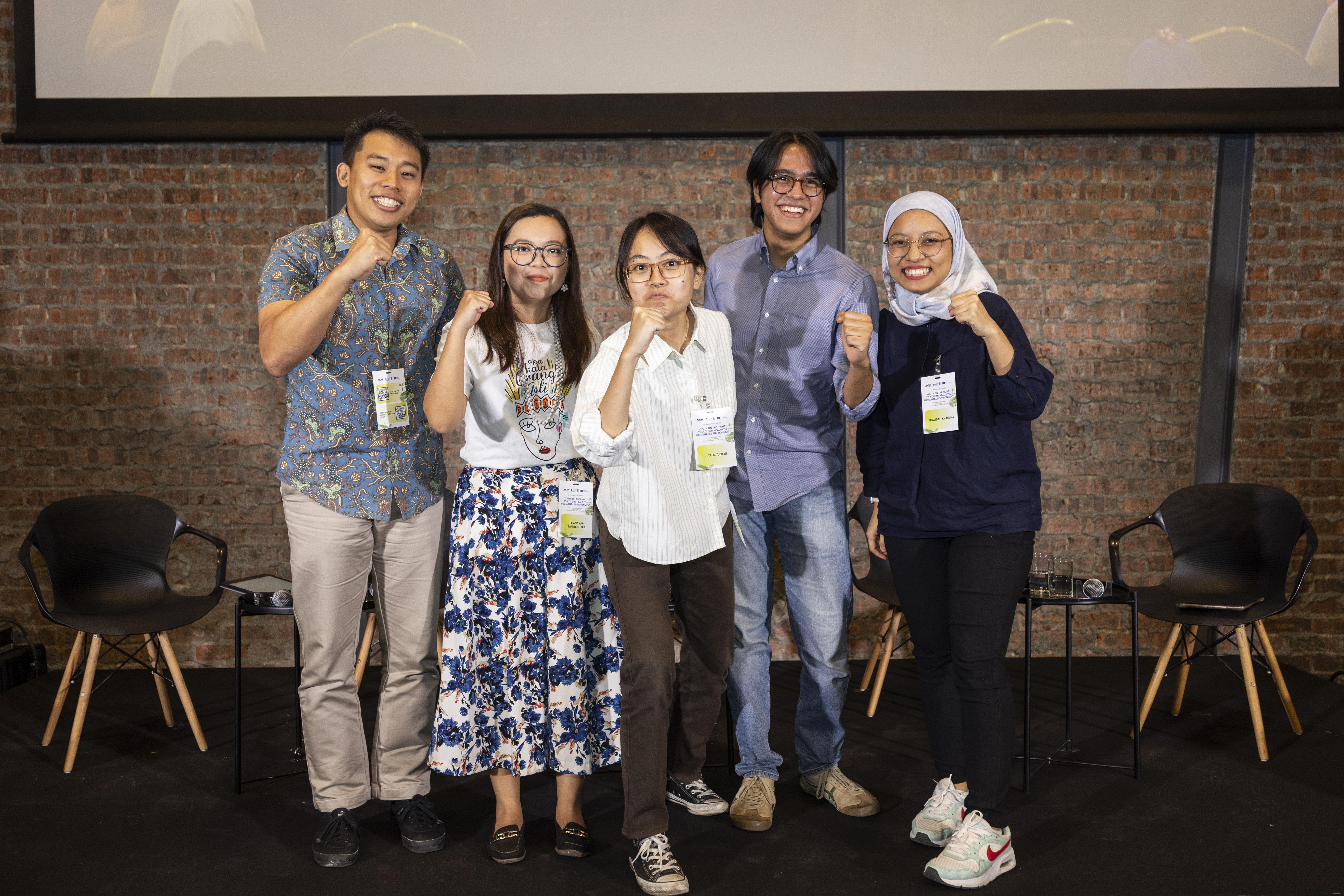 Five people standing together, smiling and making hand gestures, in a studio setting with brick walls.