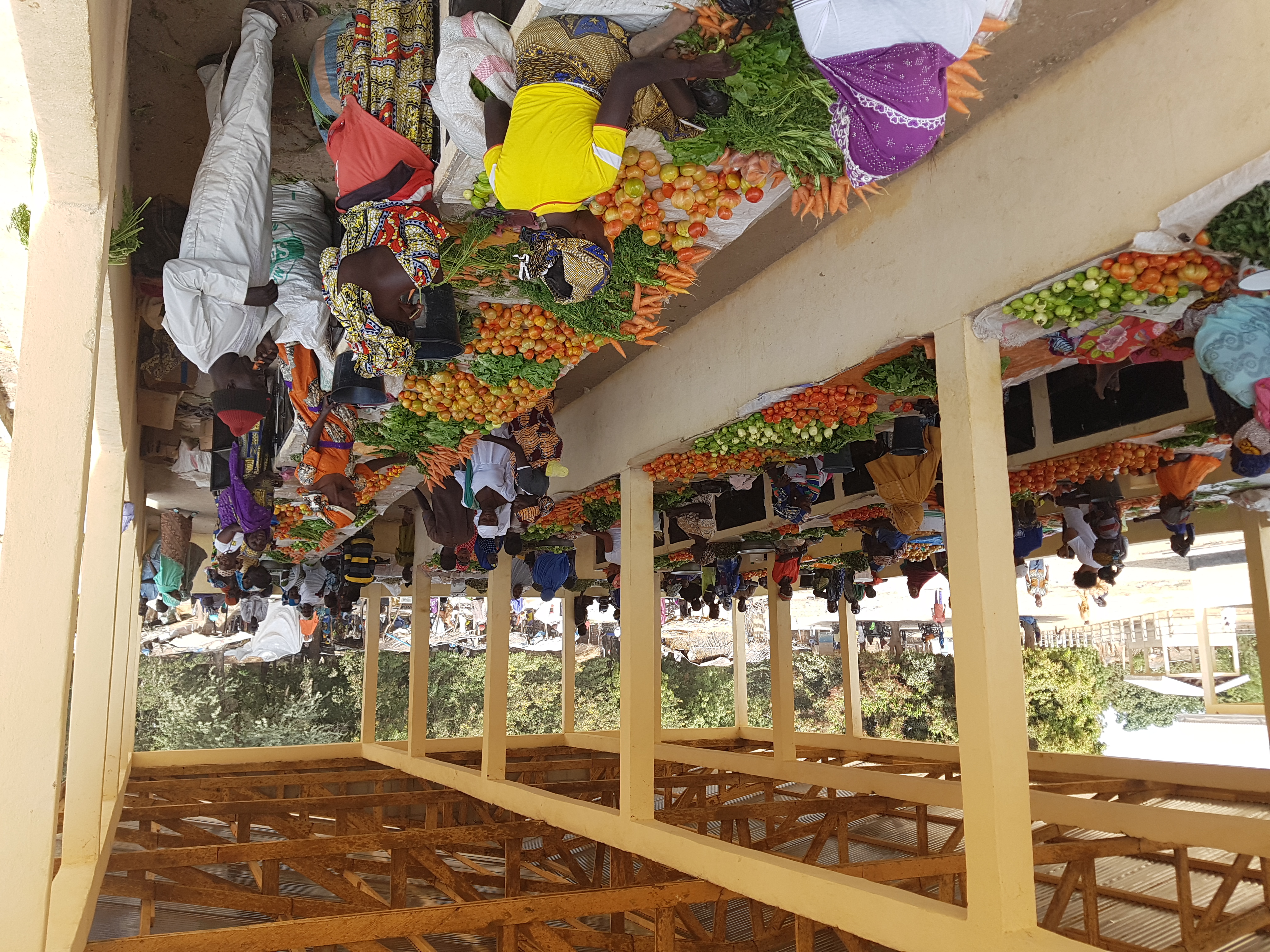A bustling outdoor market with people selling colorful vegetables and fruits under a wooden structure.