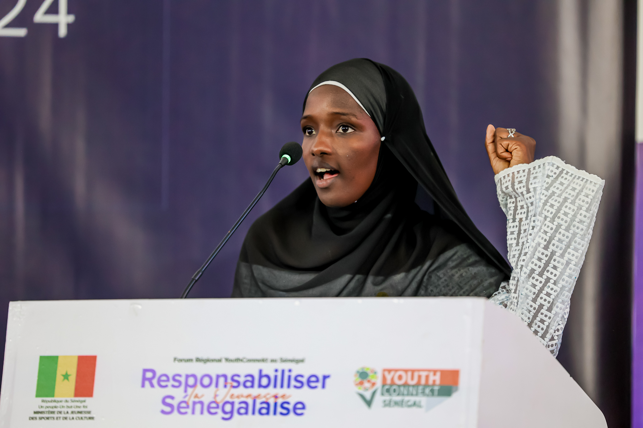 A young woman in a hijab passionately speaks at a podium during an event.