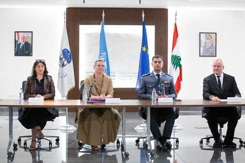 Four individuals seated at a conference table with flags and portraits in the background.