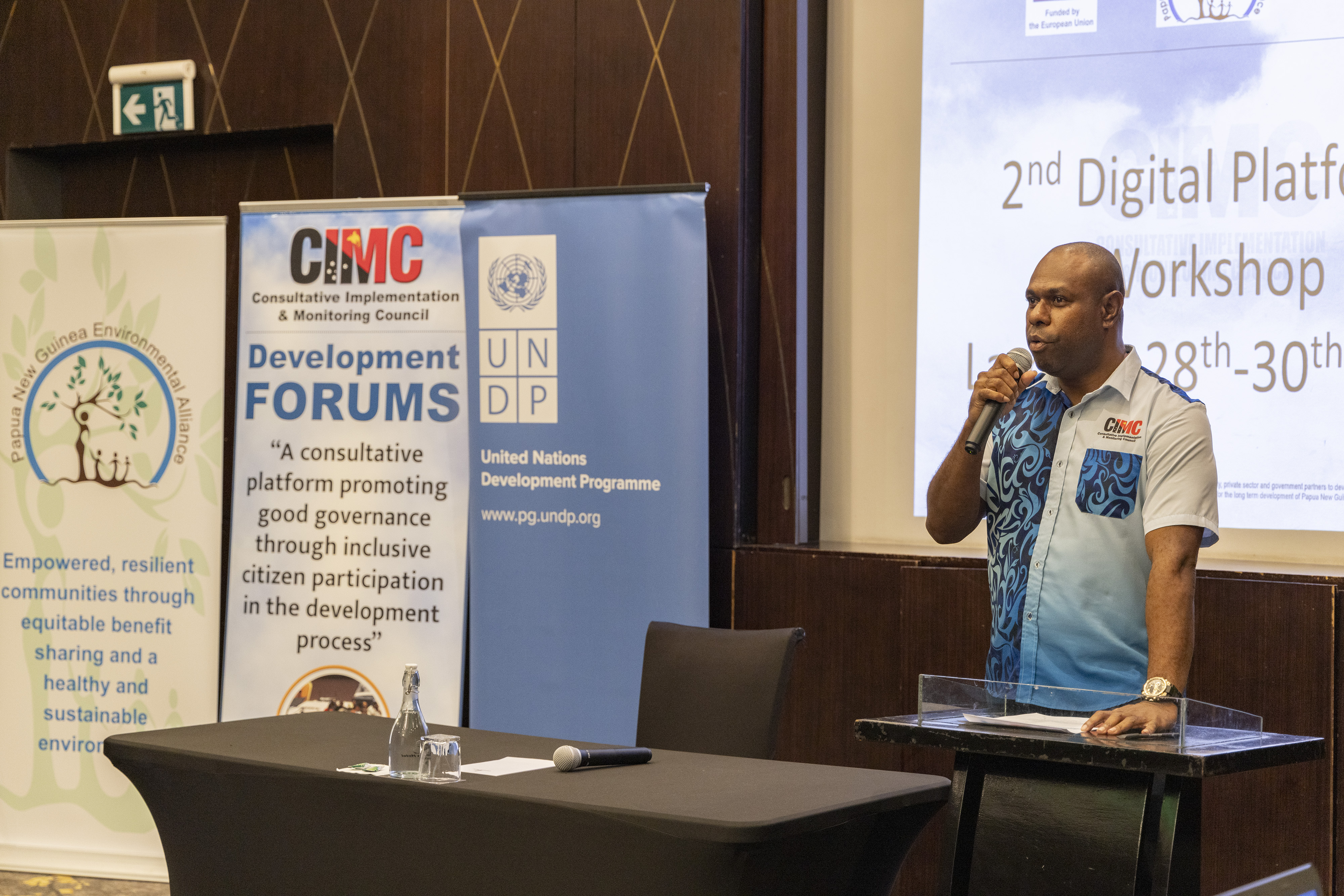 A man in a blue shirt speaks at a workshop, with banners in the background.