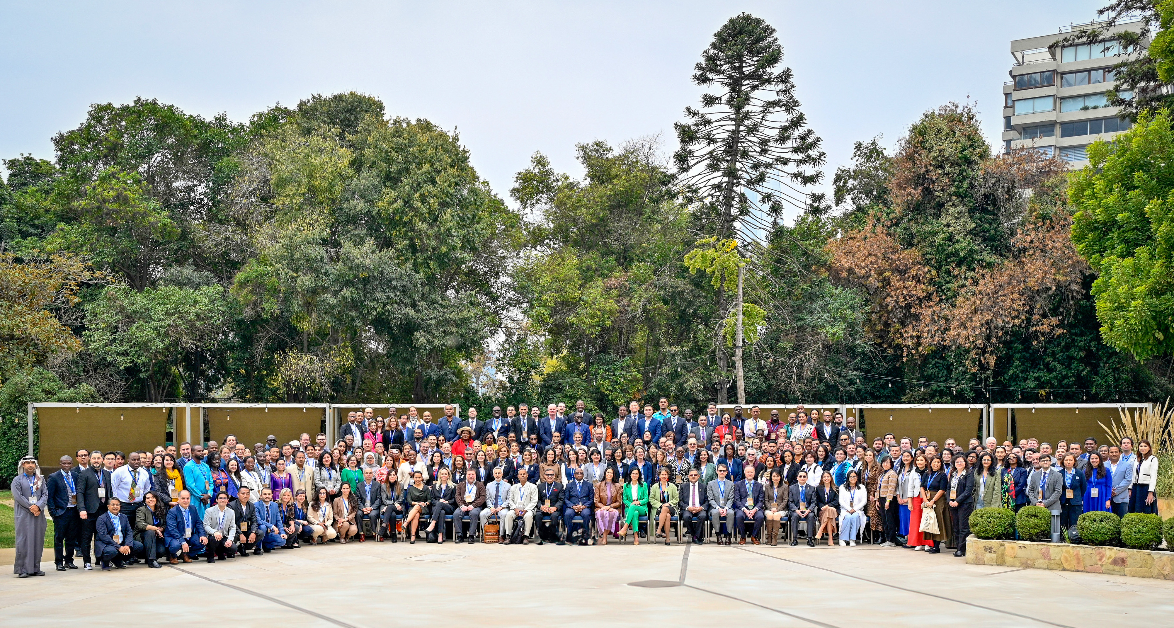 A large group of diverse people standing together outdoors, surrounded by greenery.