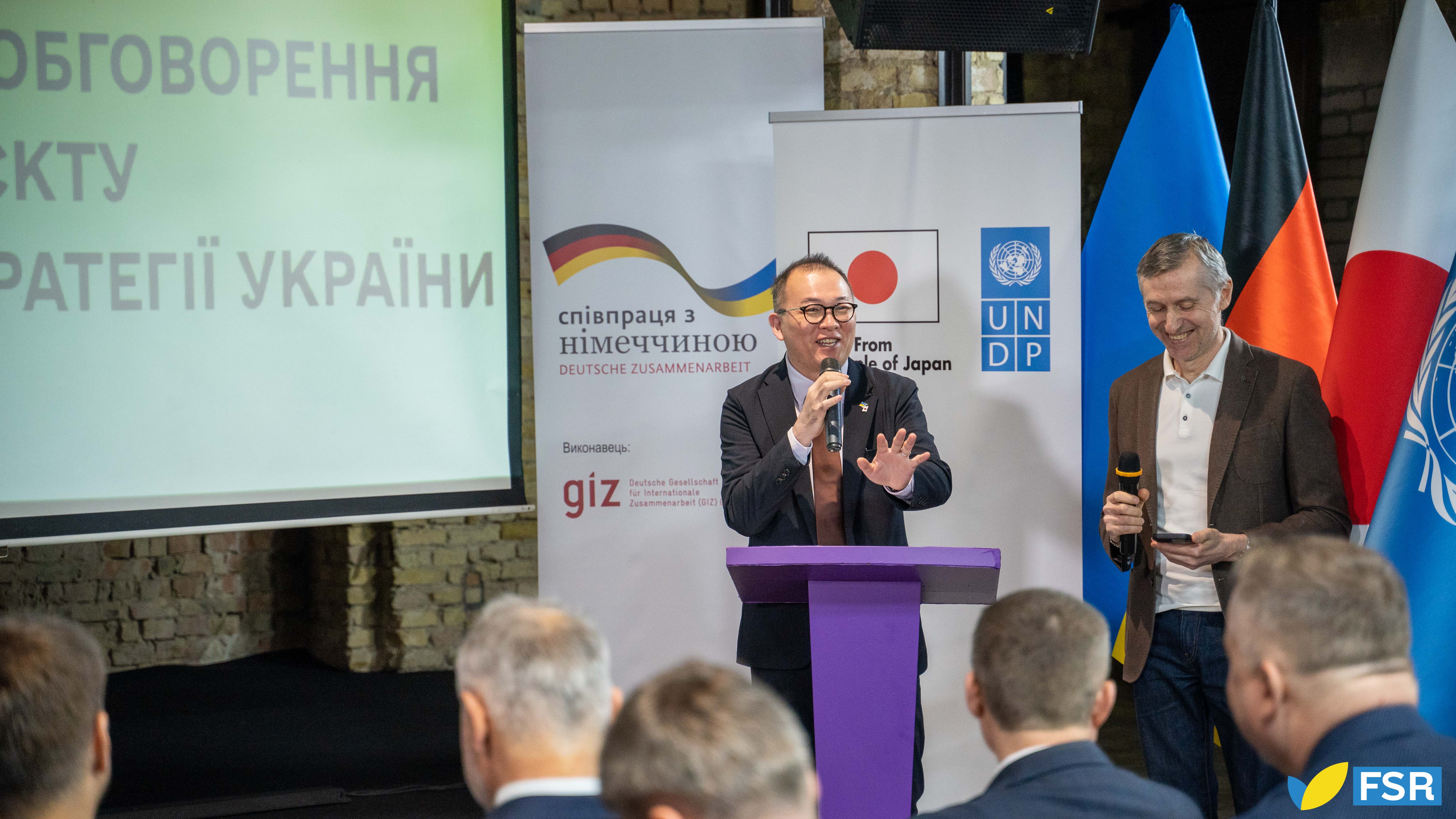 Two men speak at a conference with flags in the background and a presentation screen.