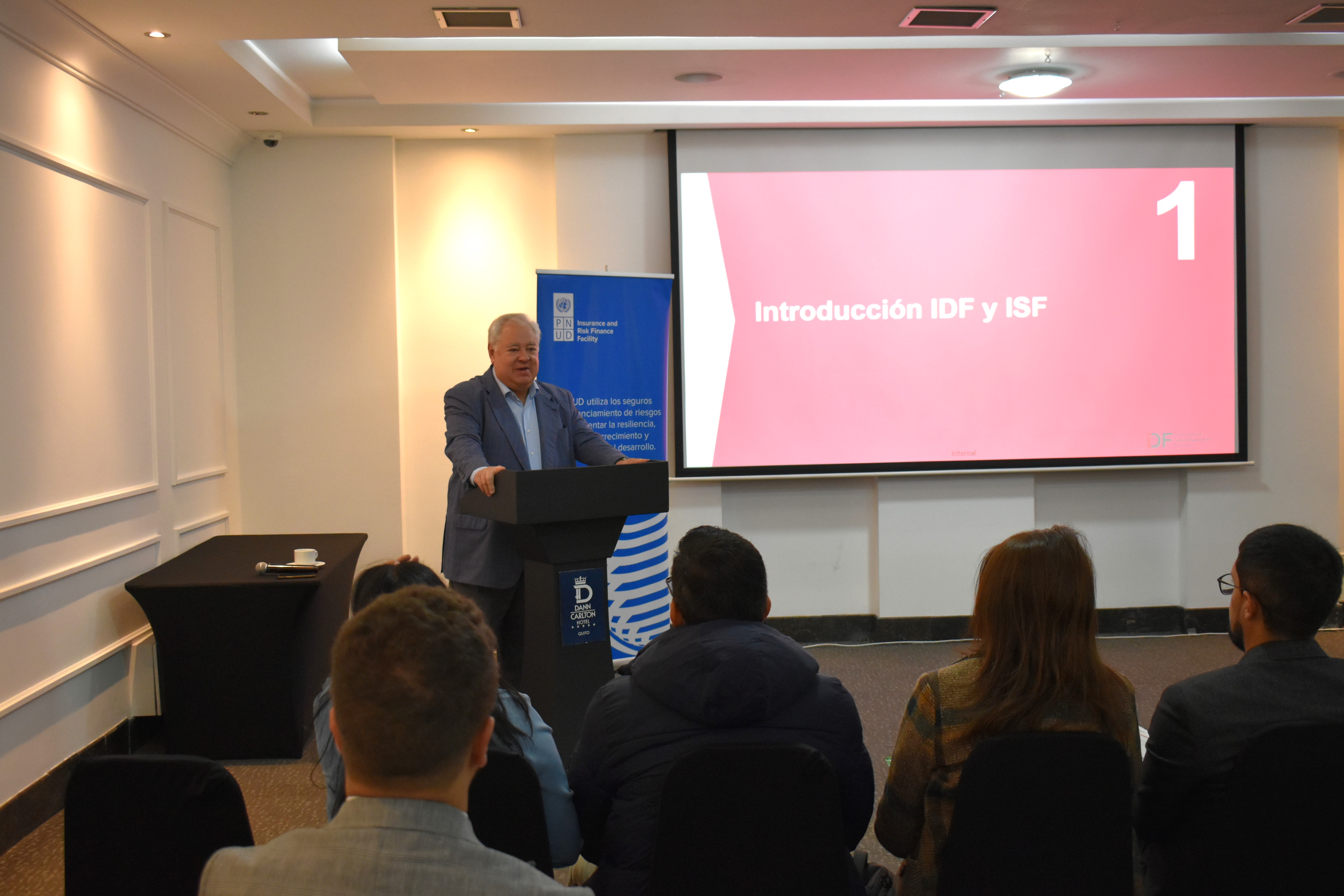 A speaker stands at a podium in front of a screen displaying "1. Introducción GIF y IBF," with an audience facing him.