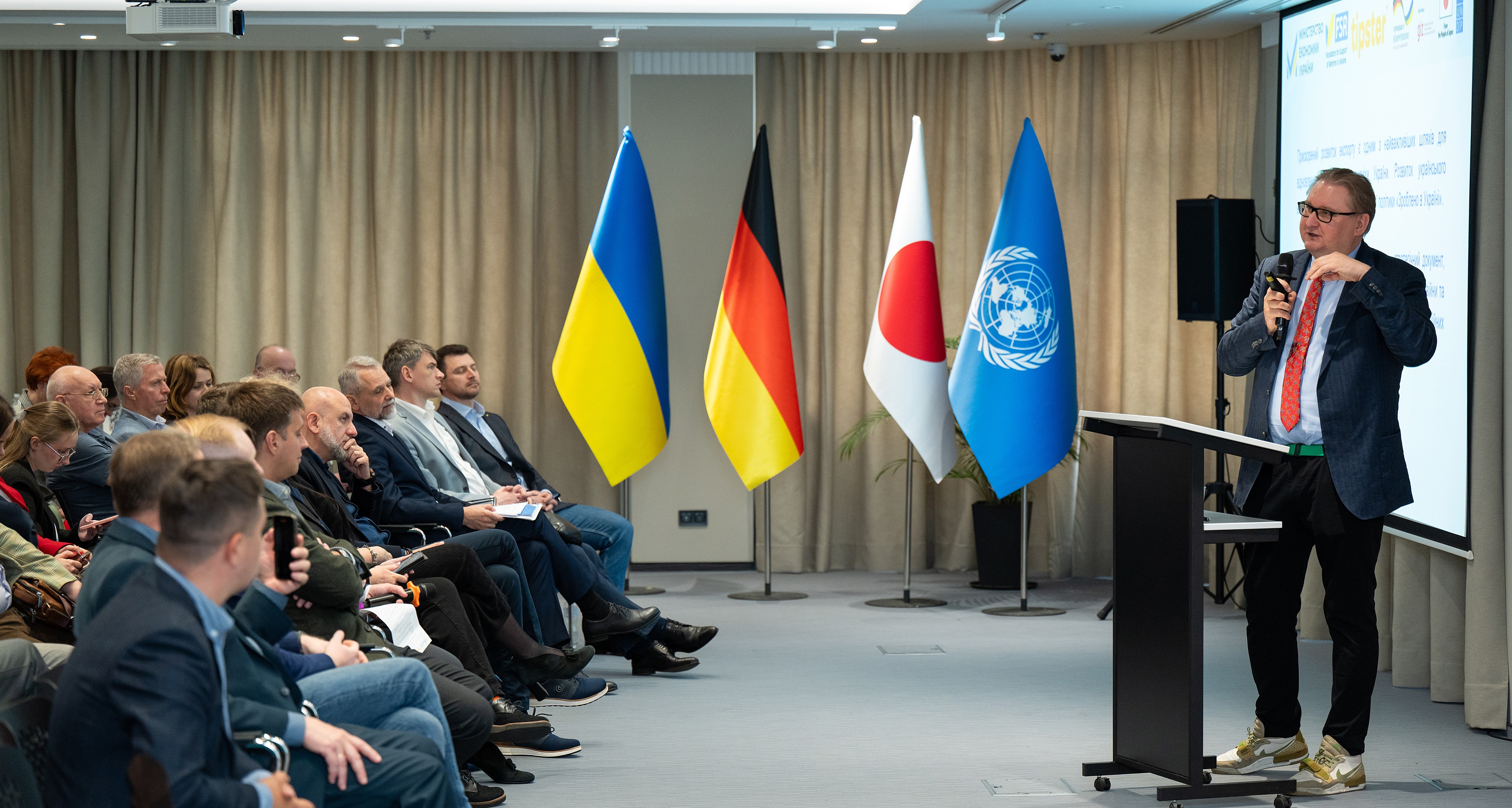 A speaker addresses an audience, with flags of Ukraine, Germany, Japan, and the UN displayed.
