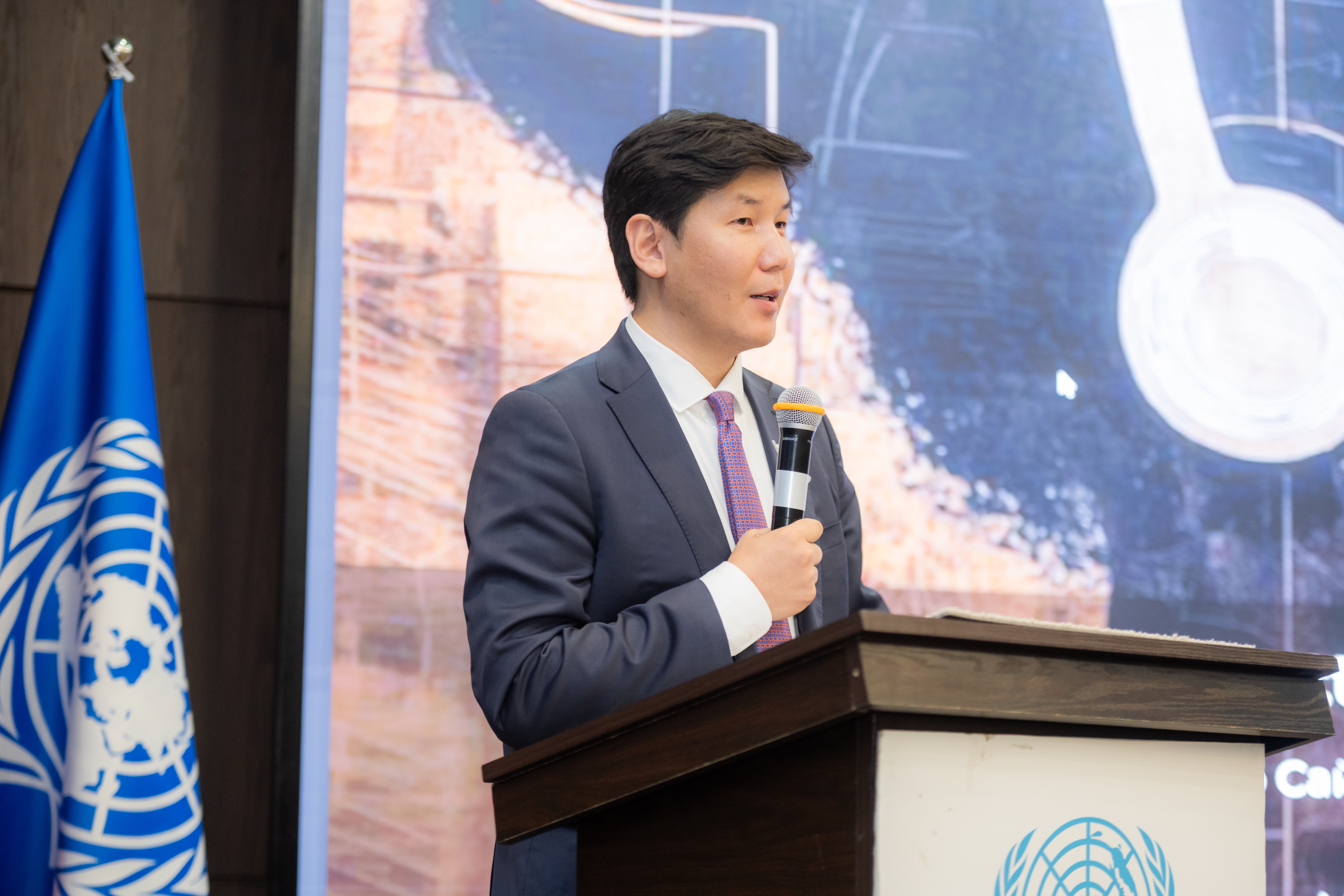 A young man in a suit speaks at a podium with a United Nations flag behind him.