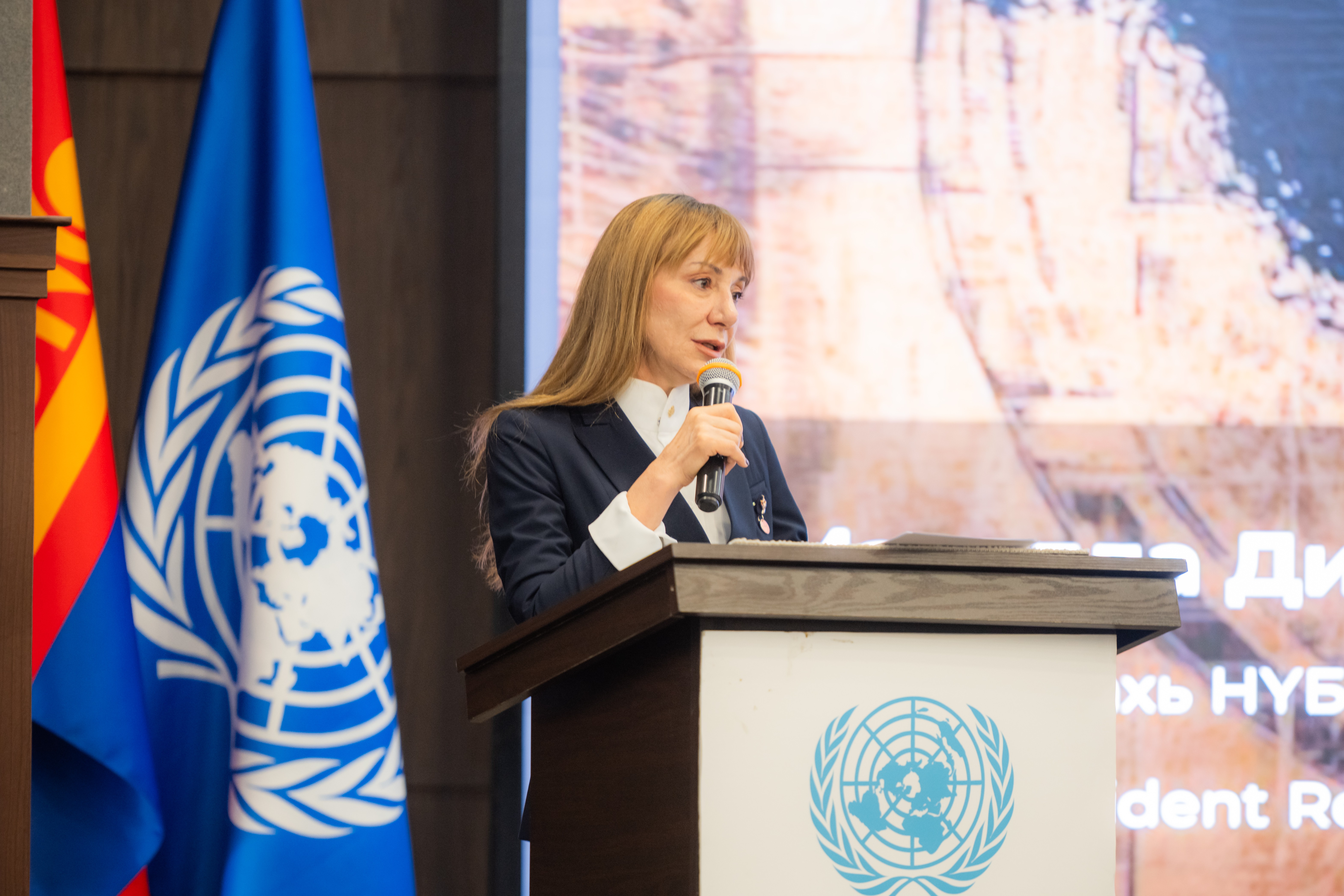 A woman speaks at a podium with United Nations branding, flanked by flags.