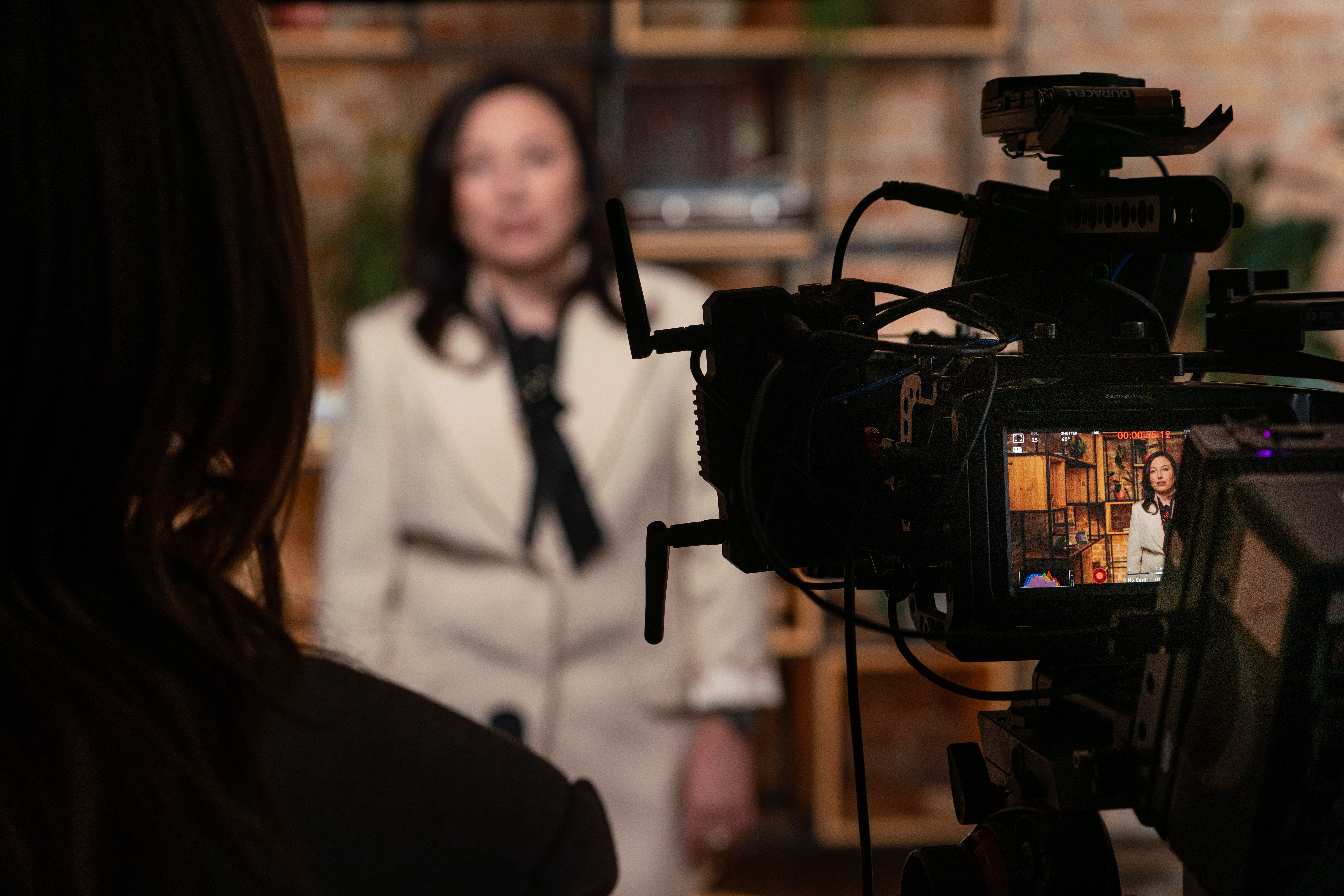 A woman in a cream suit stands in front of a camera, with shelves in the background.