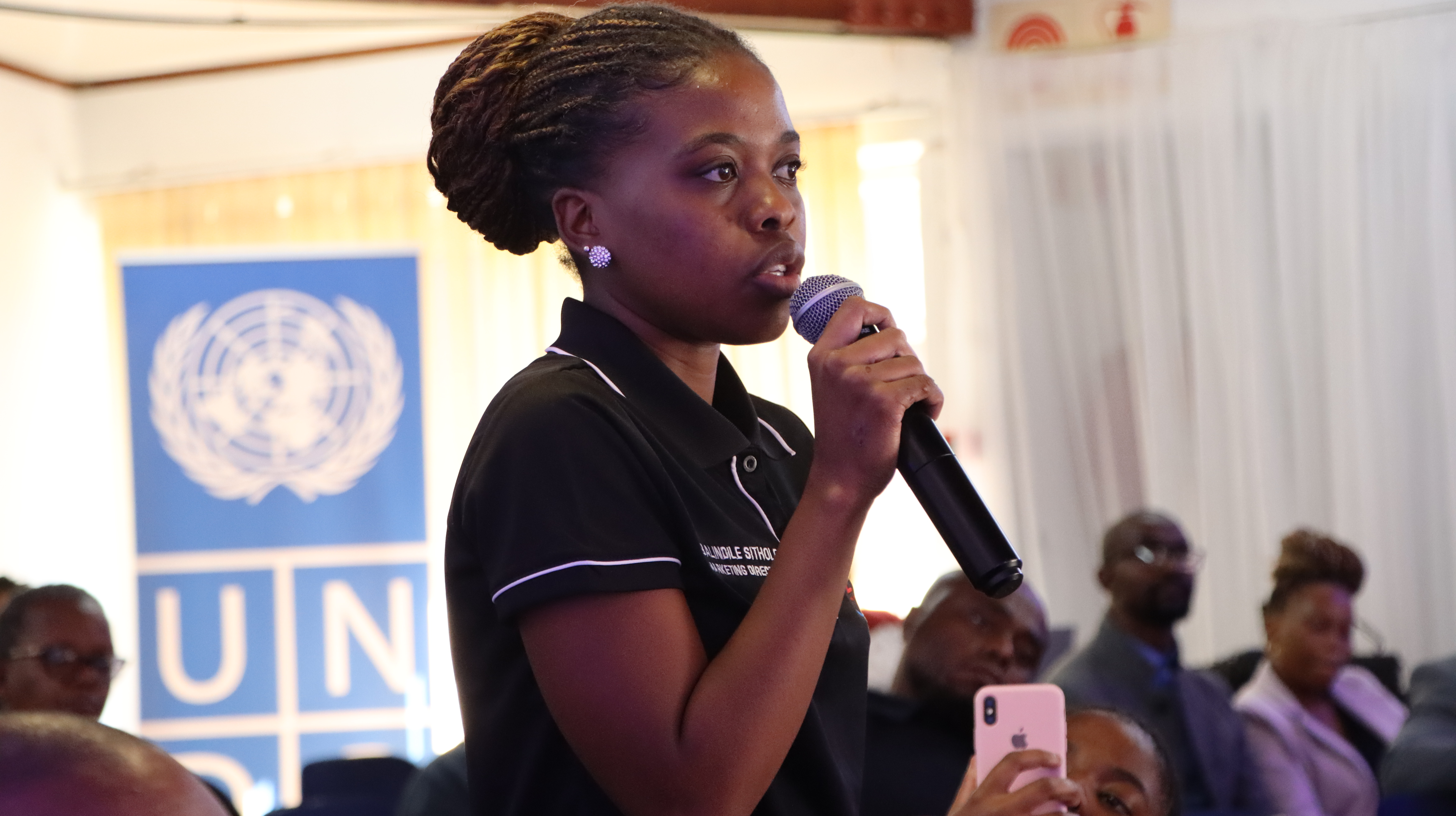 A woman speaking into a microphone with a UN banner in the background.