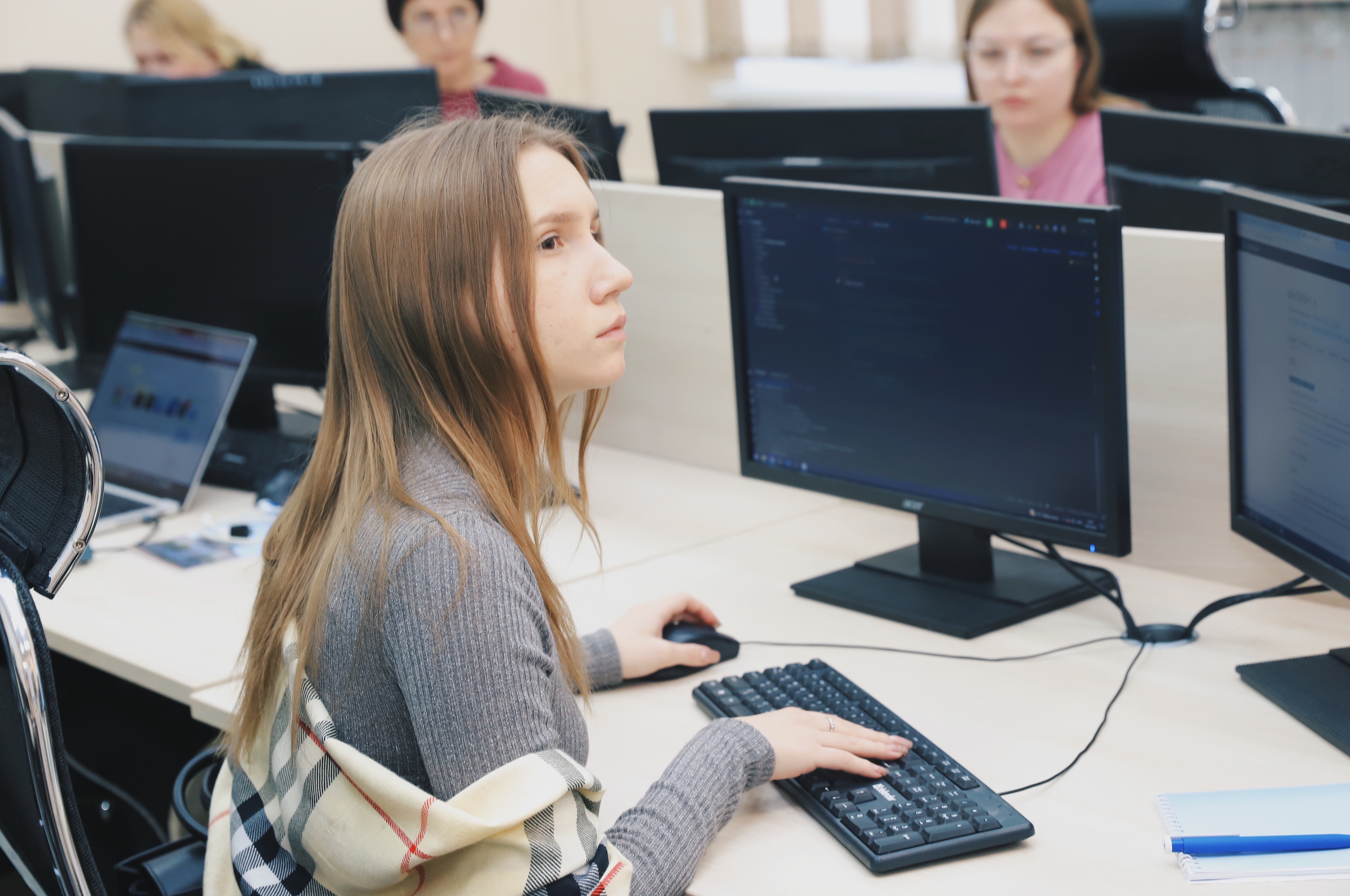 A young woman focused on her computer screen, with other students in the background.