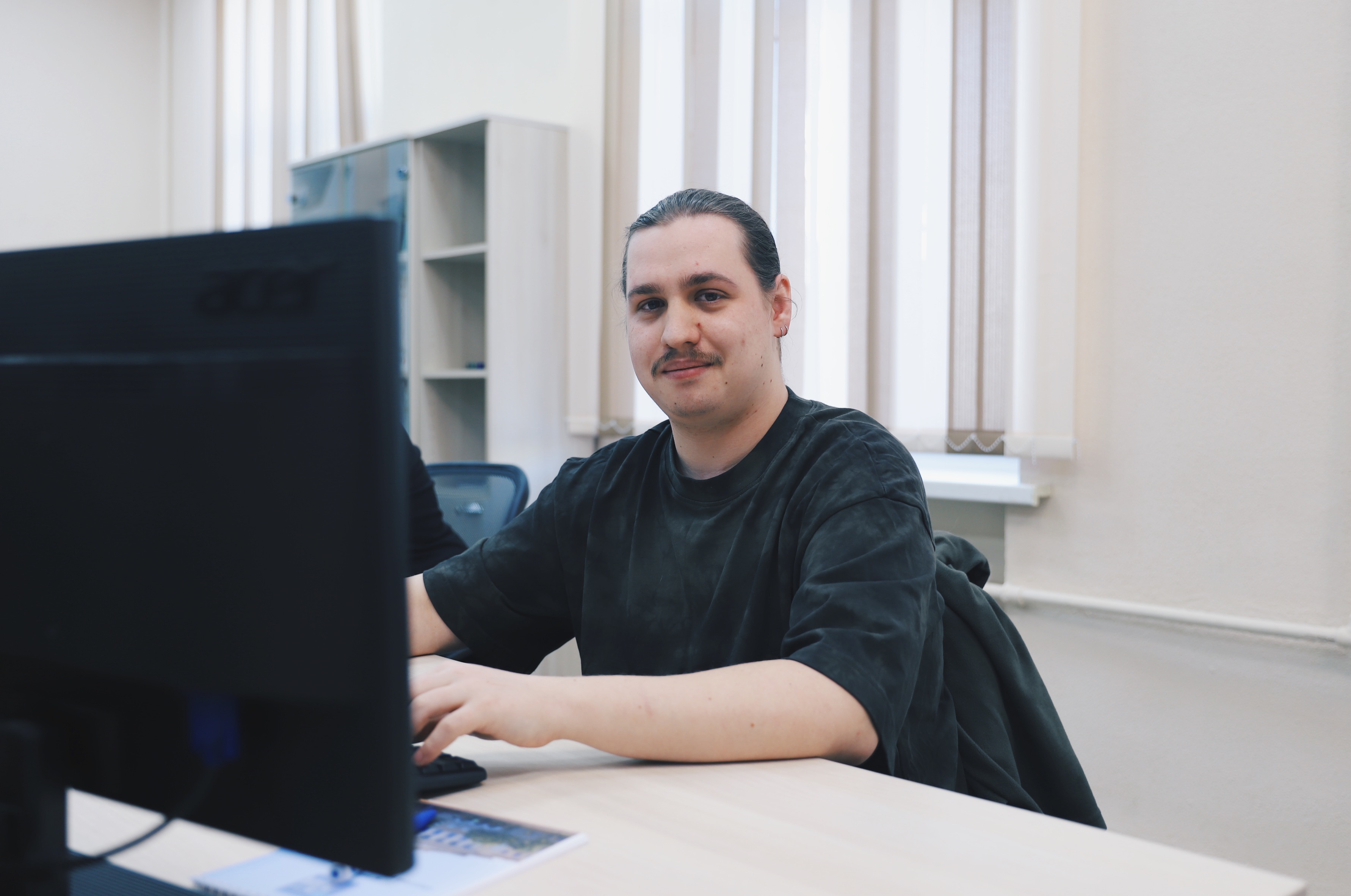 Man with a mustache sitting at a desk, smiling at a computer monitor in a bright room.