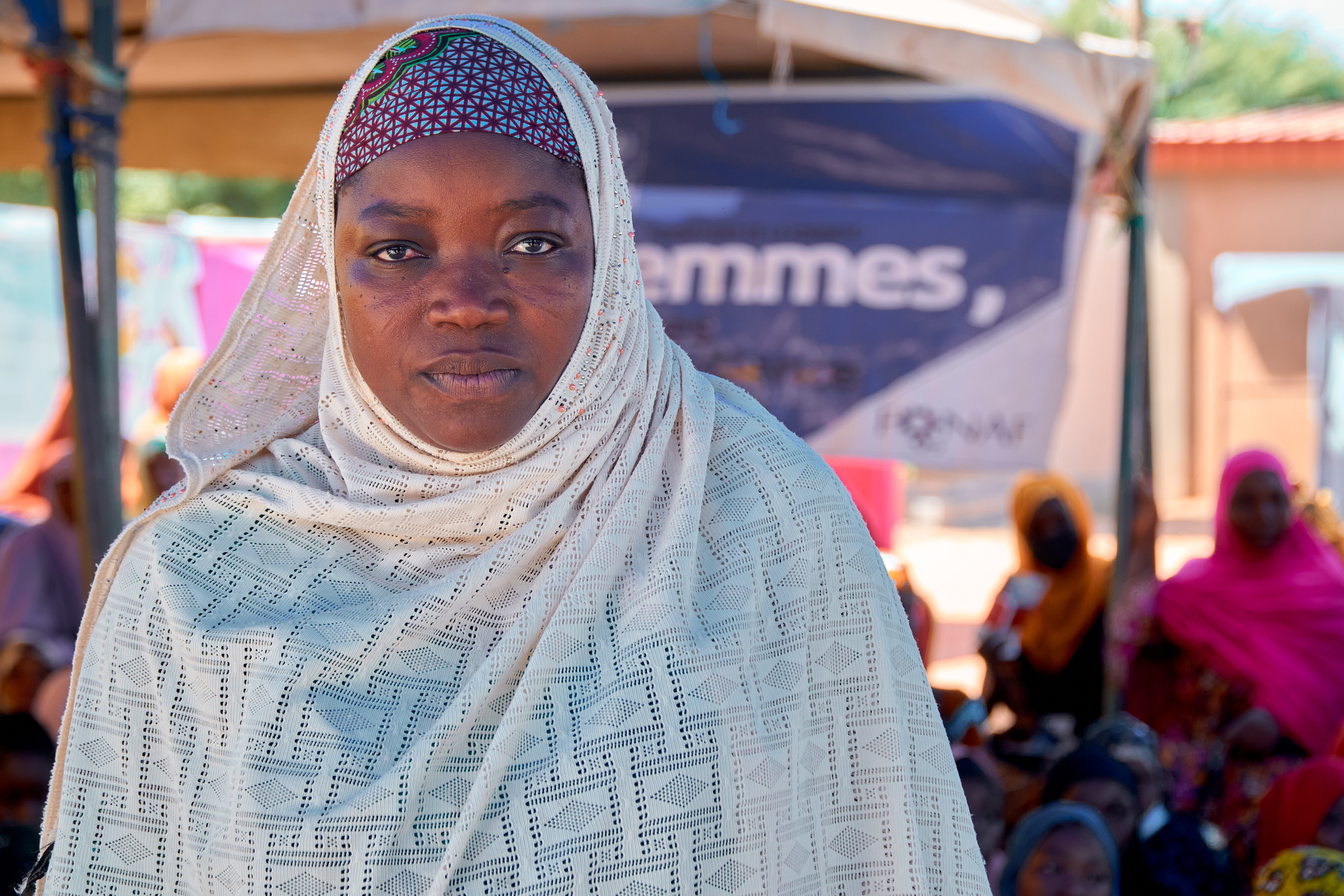 A woman in a white headscarf stands in front of a gathering, looking directly at the camera.