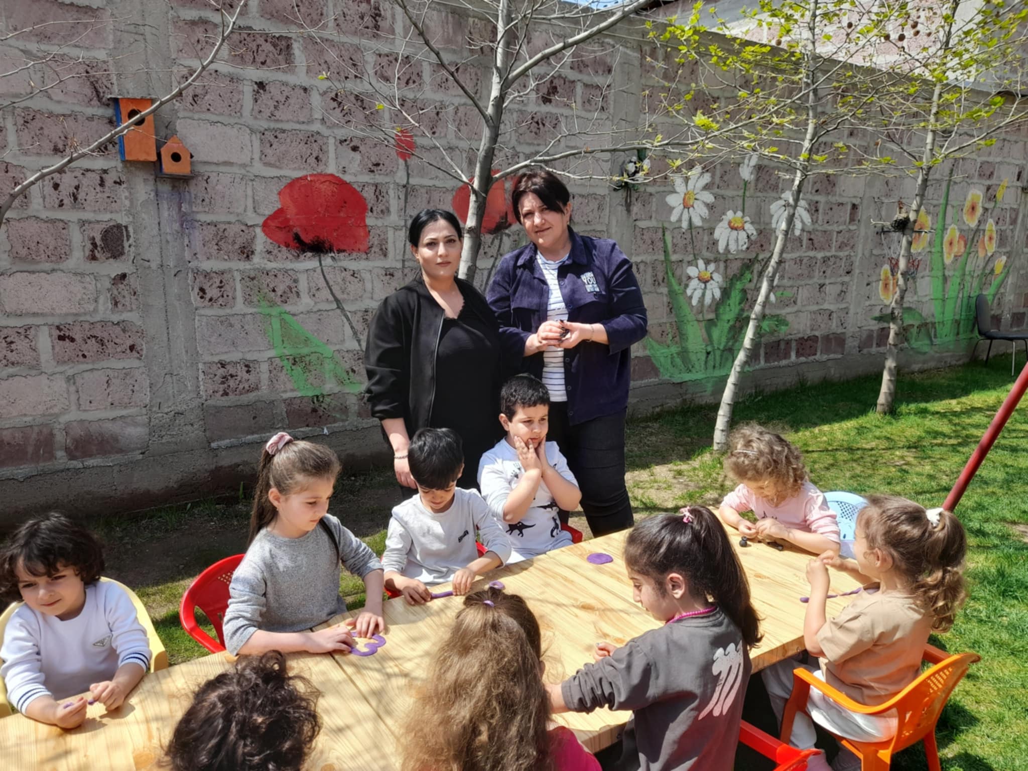 Children and adults engage in activities at a table in a colorful outdoor setting.